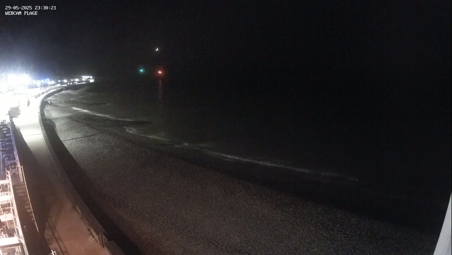 A dark, nighttime view of a pebble beach with gently lapping waves under a crescent moon.