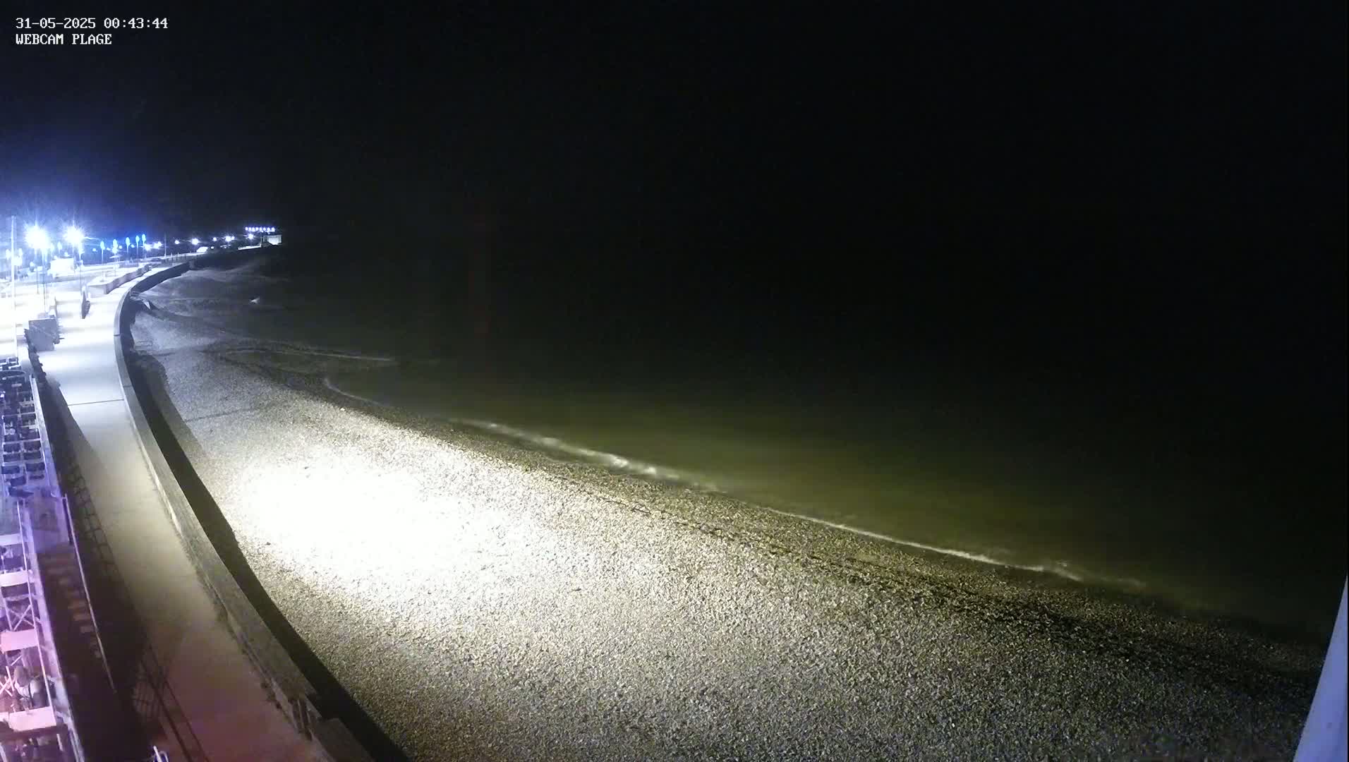 A dark, nighttime beach scene shows a pebble shore gently lapped by waves under artificial light from street lamps.
