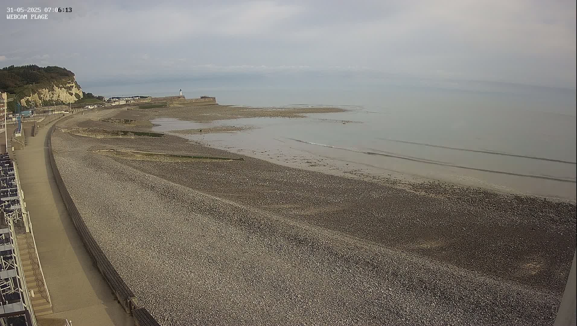 A pebble beach stretches from a paved walkway towards a calm sea under an overcast sky, with a cliff and lighthouse visible in the distance.