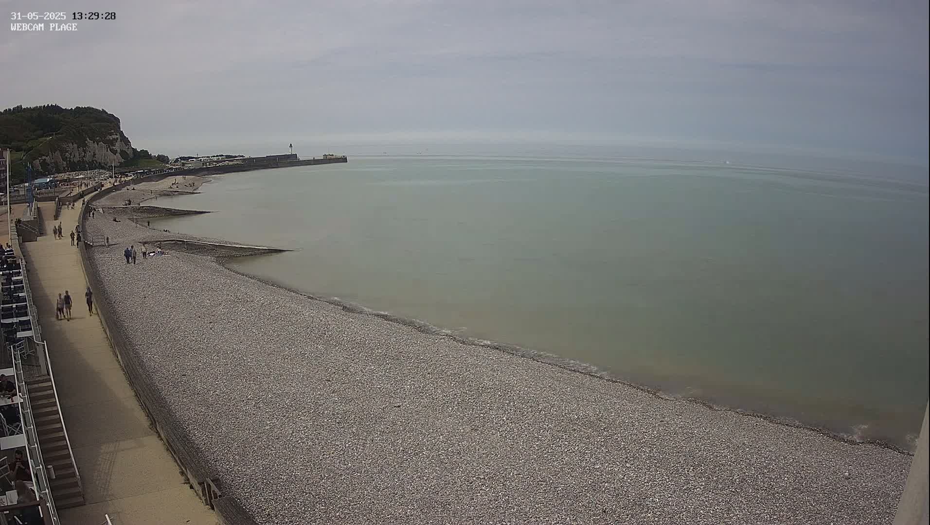 A pebble beach stretches along a calm, gray-green sea under an overcast sky, with people walking along a promenade and some relaxing on the shore.