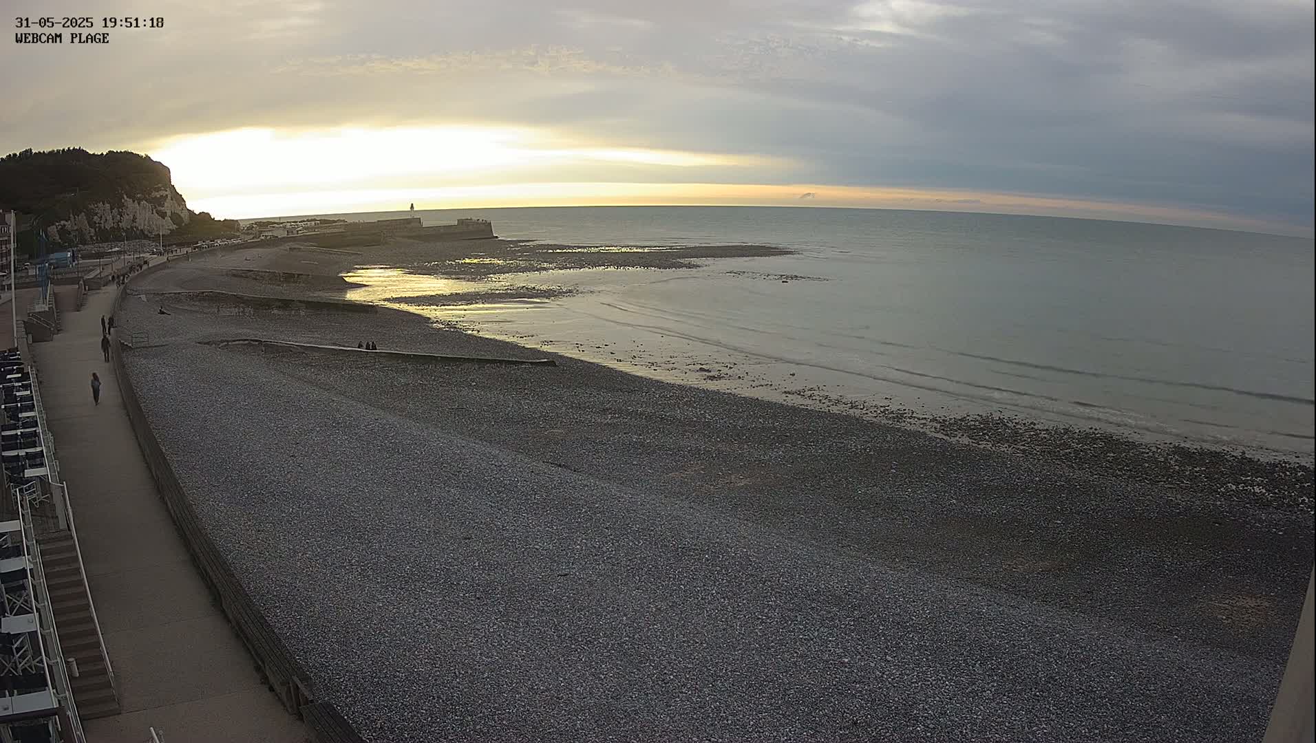 A pebble beach with a few people walking along a promenade, under a partly cloudy sunset sky.