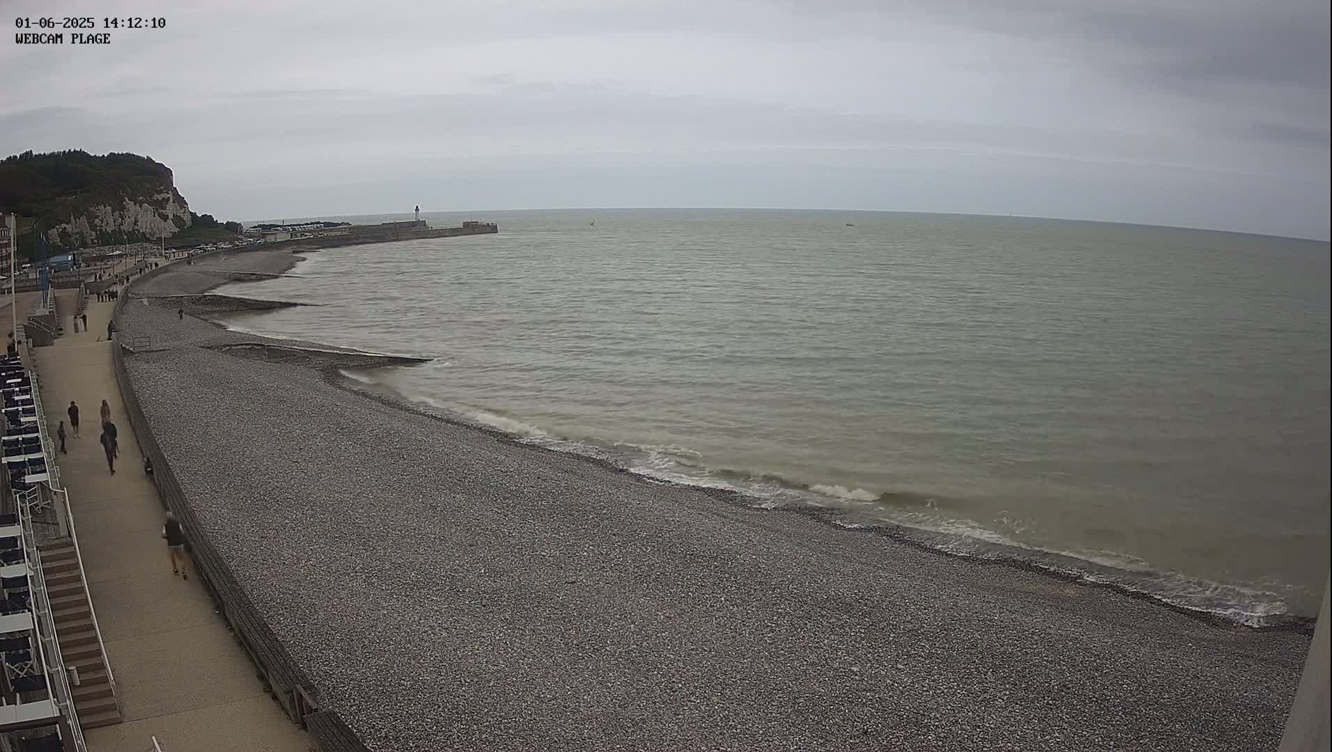 A pebble beach stretches along a calm, gray sea under an overcast sky, with a walkway and a few people visible along the shore.