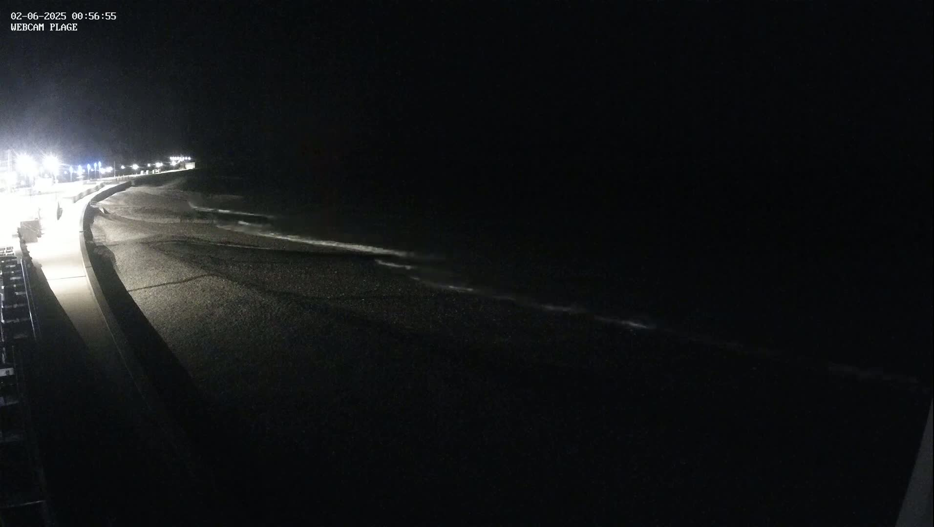 A dark, nighttime view of a pebble beach with gently lapping waves and a lit walkway along the shore.