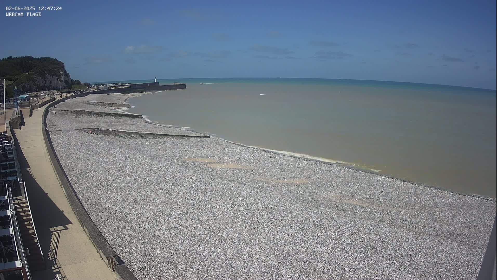 A pebble beach stretches along a calm, grayish-brown sea under a clear blue sky, with a stone seawall and a distant cliff visible.