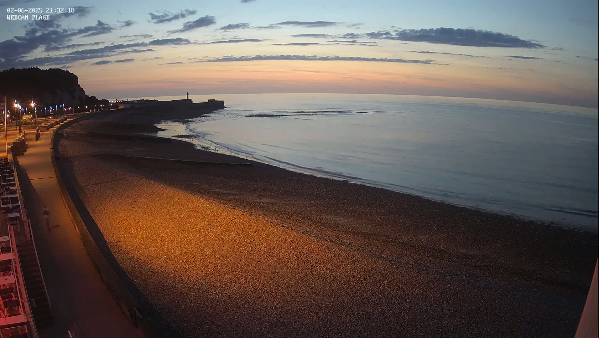 A pebble beach at dusk, with a calm sea and a promenade lit by streetlights, under a mostly clear sky.