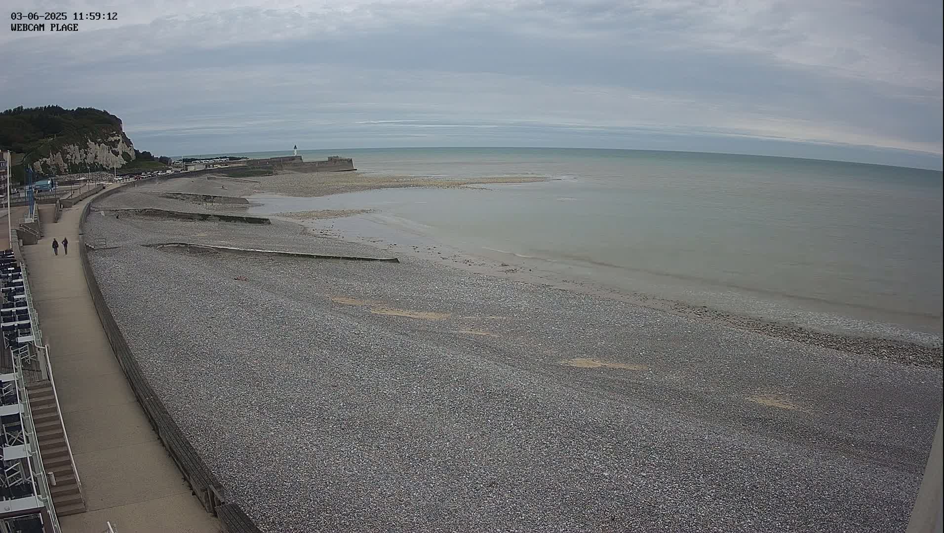 A pebble beach stretches along a calm, gray sea under an overcast sky, with a walkway and buildings visible in the background.