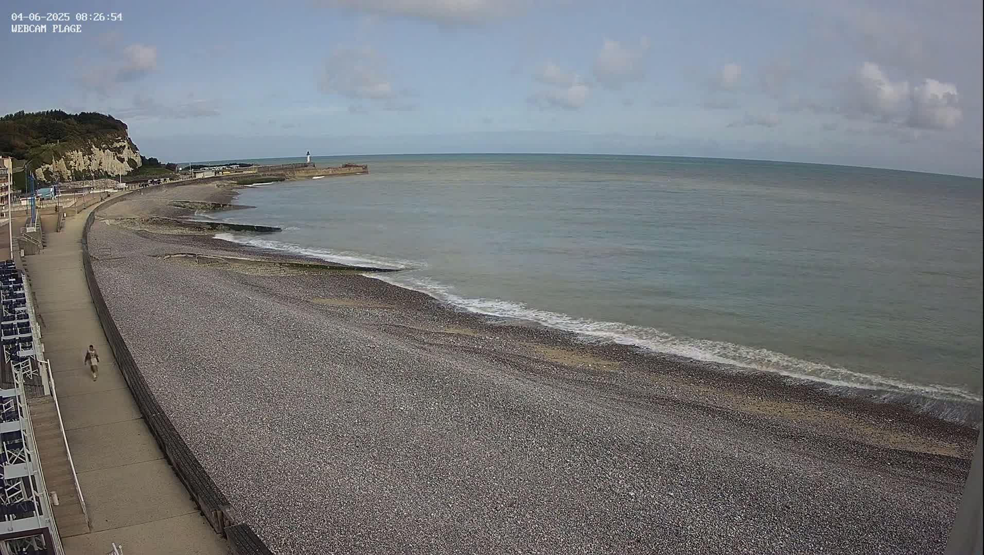 A pebble beach stretches along a calm, grey-blue sea under a partly cloudy sky, with a person walking along a paved walkway next to it and a cliff visible in the distance.