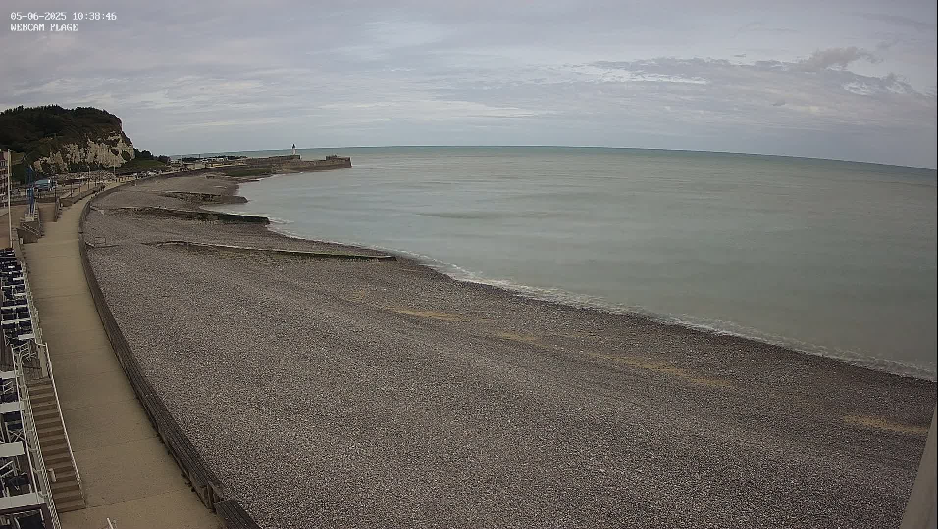 A pebble beach stretches along a calm, gray sea under an overcast sky, with a walkway and white cliffs visible in the background.