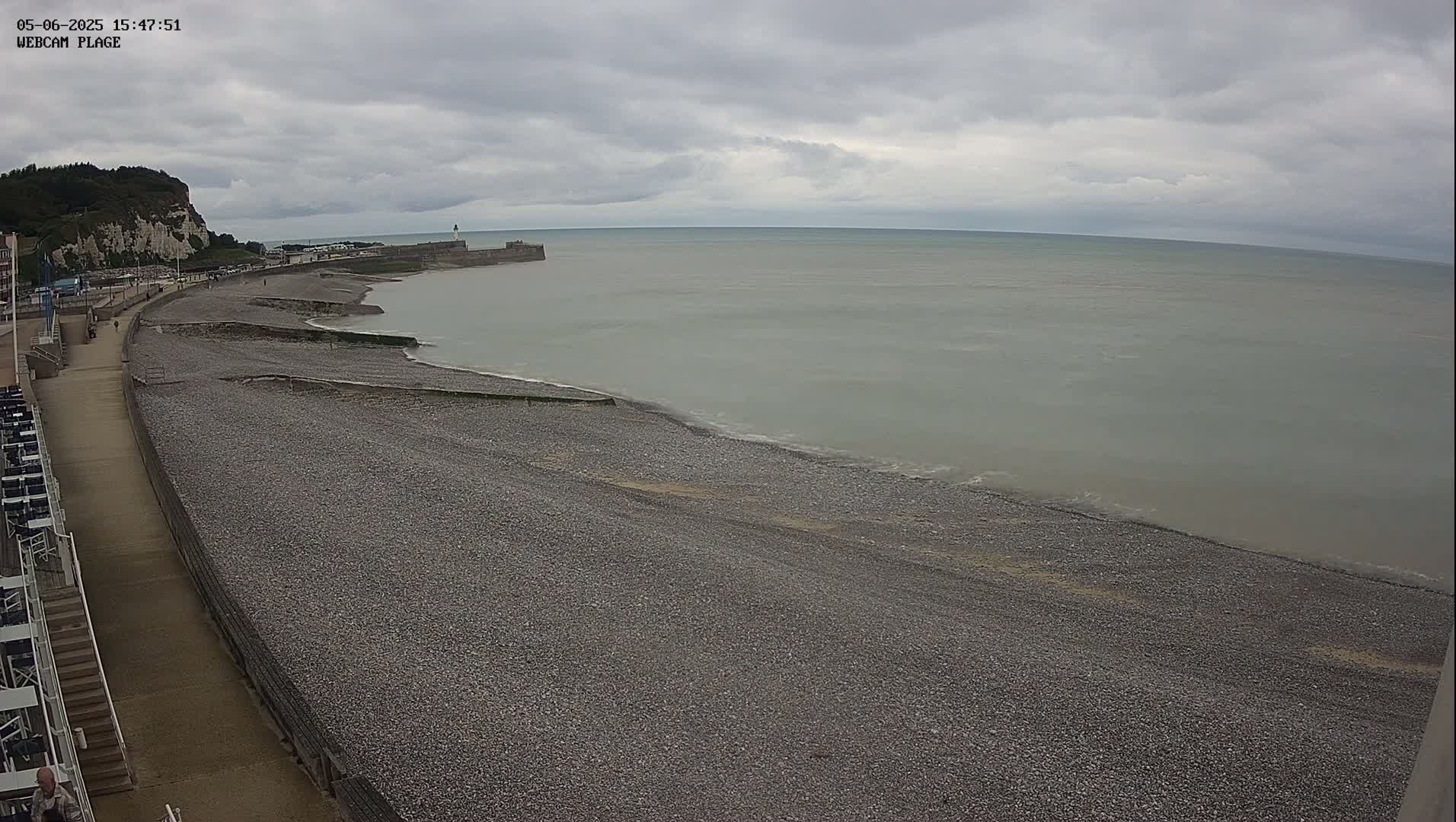 A pebble beach stretches along a calm, gray sea under an overcast sky, with a walkway and white cliffs visible in the background.
