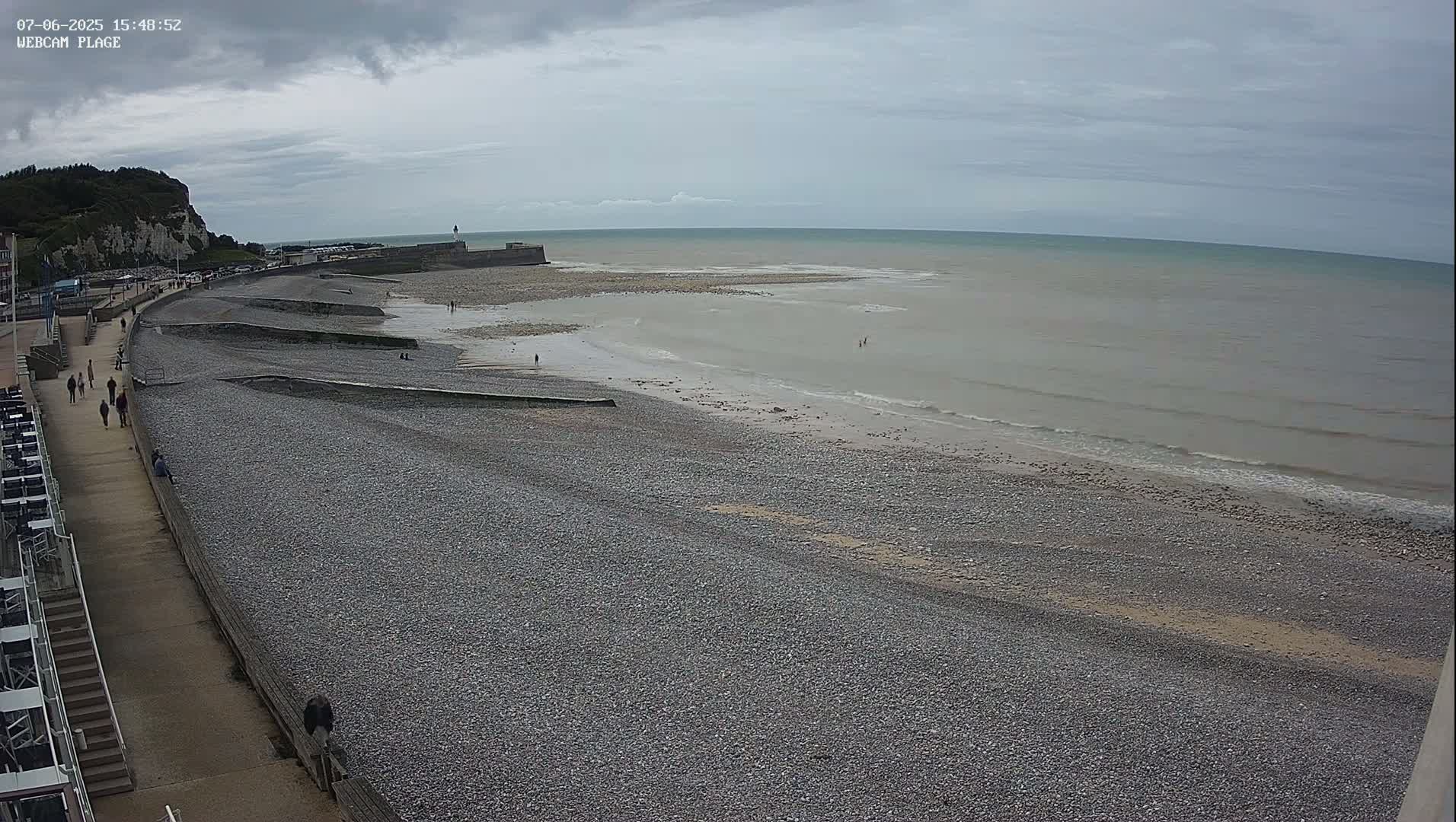 A pebble beach stretches along a calm, gray-toned sea under an overcast sky, with a few people walking on the beach and promenade.