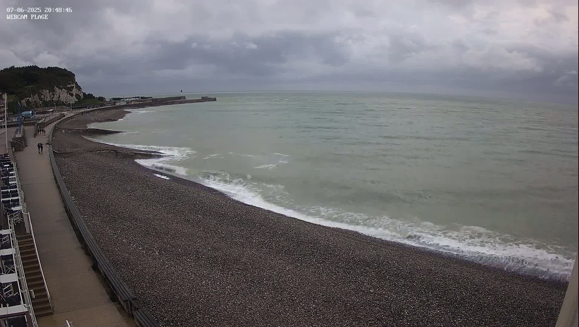 A pebble beach with gentle waves laps the shore under an overcast sky, with a paved walkway and distant cliffs visible.