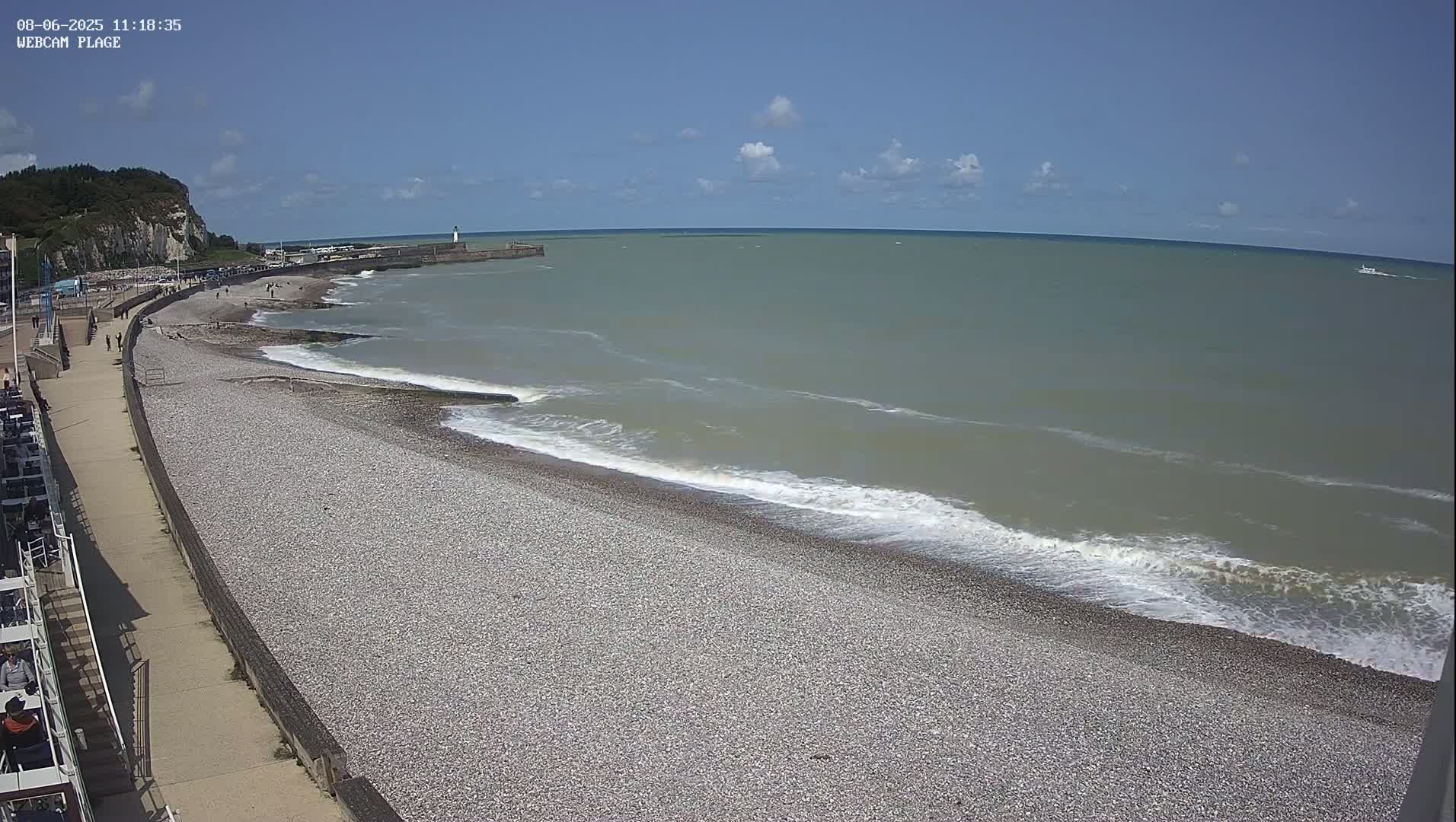 A pebble beach with gentle waves under a mostly clear blue sky stretches out to a calm sea; a few people are walking along the beach and a promenade.
