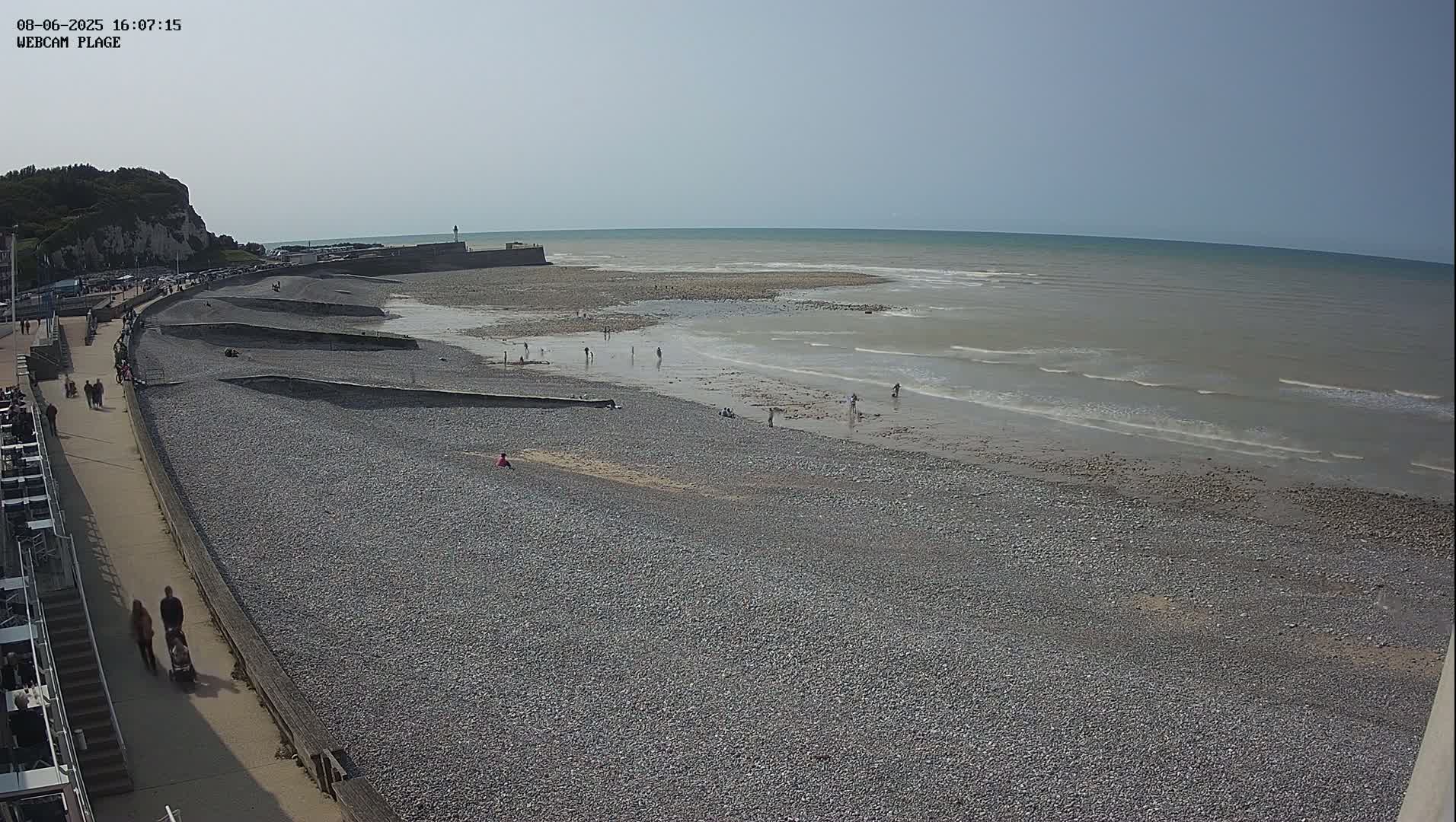 A pebble beach with a few people on it stretches out to a calm sea under a bright, sunny sky.