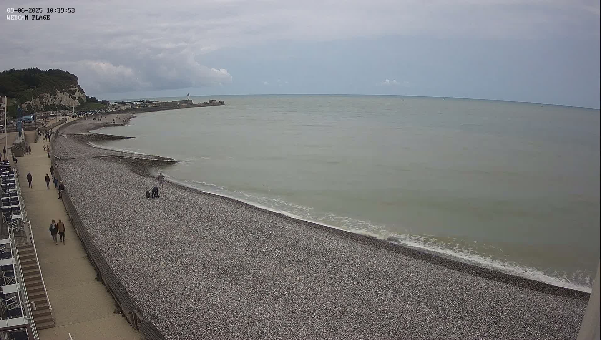 A pebble beach with a few people walking along a paved walkway, next to calm, light-greenish water under a mostly cloudy sky.