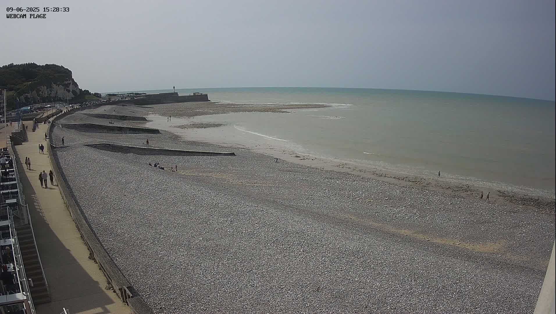 A pebble beach stretches along a calm, light-grey sea under a bright, clear sky, with people walking on the beach and a promenade.