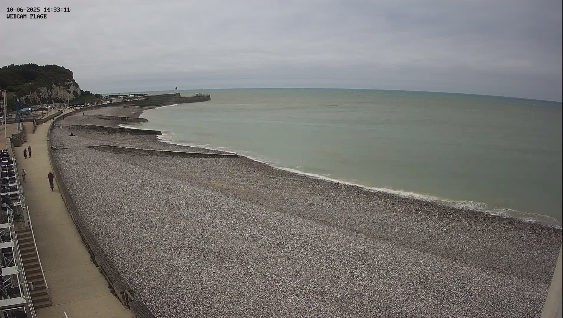 A pebble beach stretches along a calm, gray sea under an overcast sky, with a paved walkway and a few people visible.