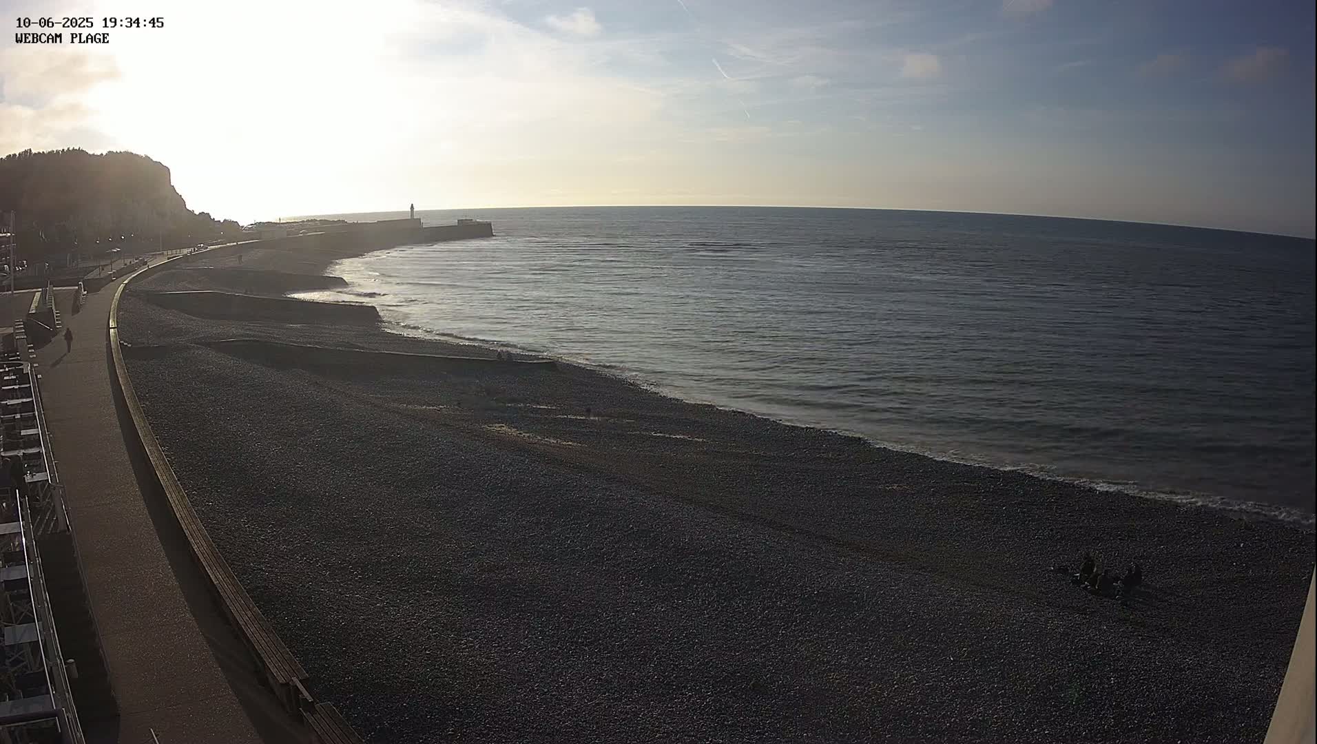 A dark pebble beach stretches along a calm sea under a mostly clear sky at dusk, with a paved walkway and a cliff visible in the distance.