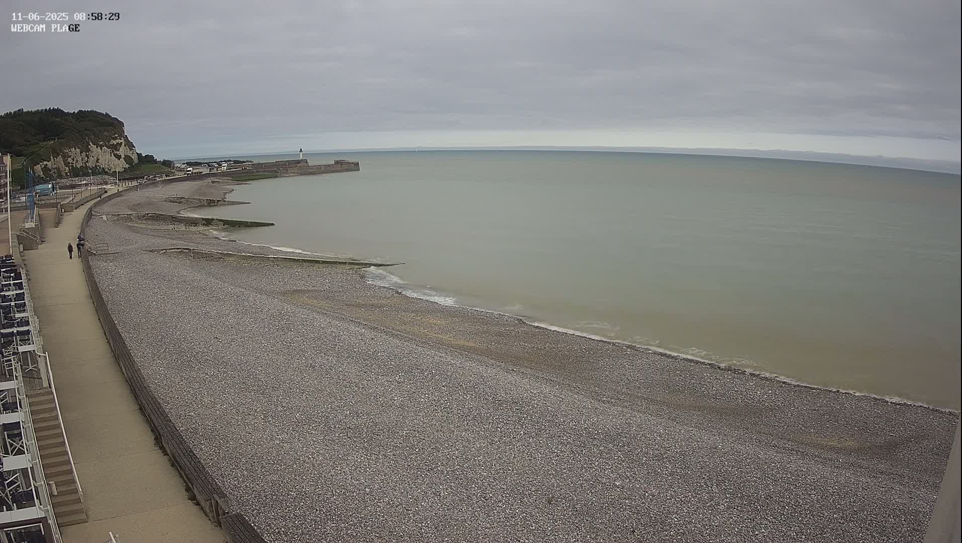 A pebble beach stretches along a calm, grey sea under an overcast sky, with a walkway and a distant cliff visible.