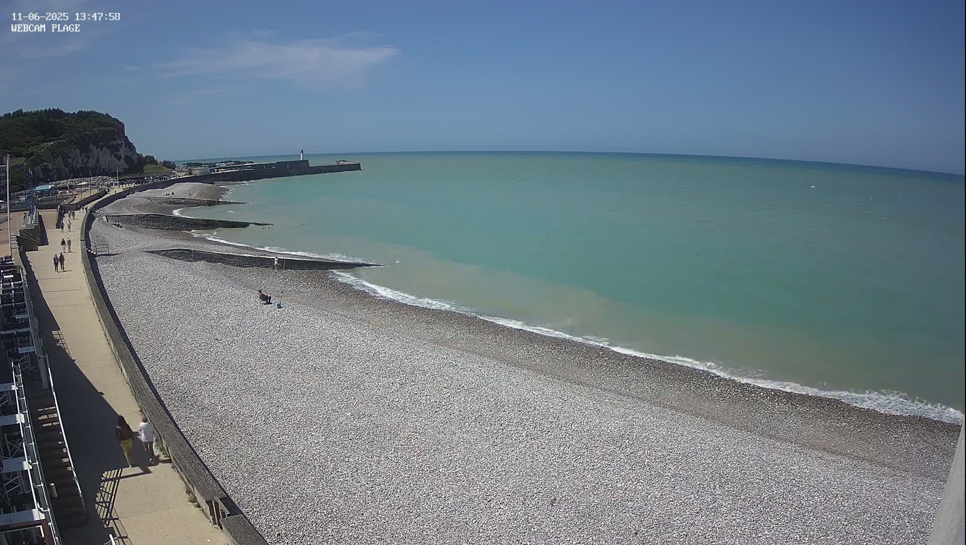 A pebble beach stretches along a calm, teal ocean under a clear blue sky, with people strolling along a promenade next to the shore and a lighthouse visible in the distance.