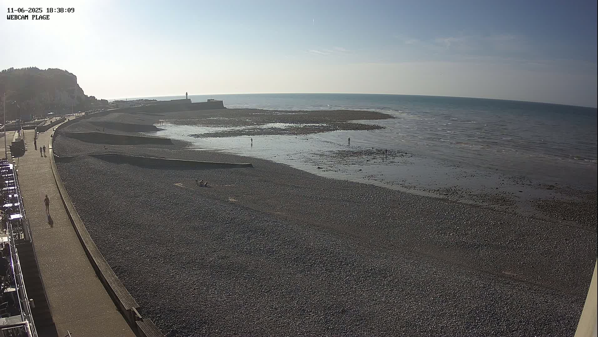 A pebbled beach stretches along a calm sea under a clear, sunny sky, with a paved walkway and a few people visible.