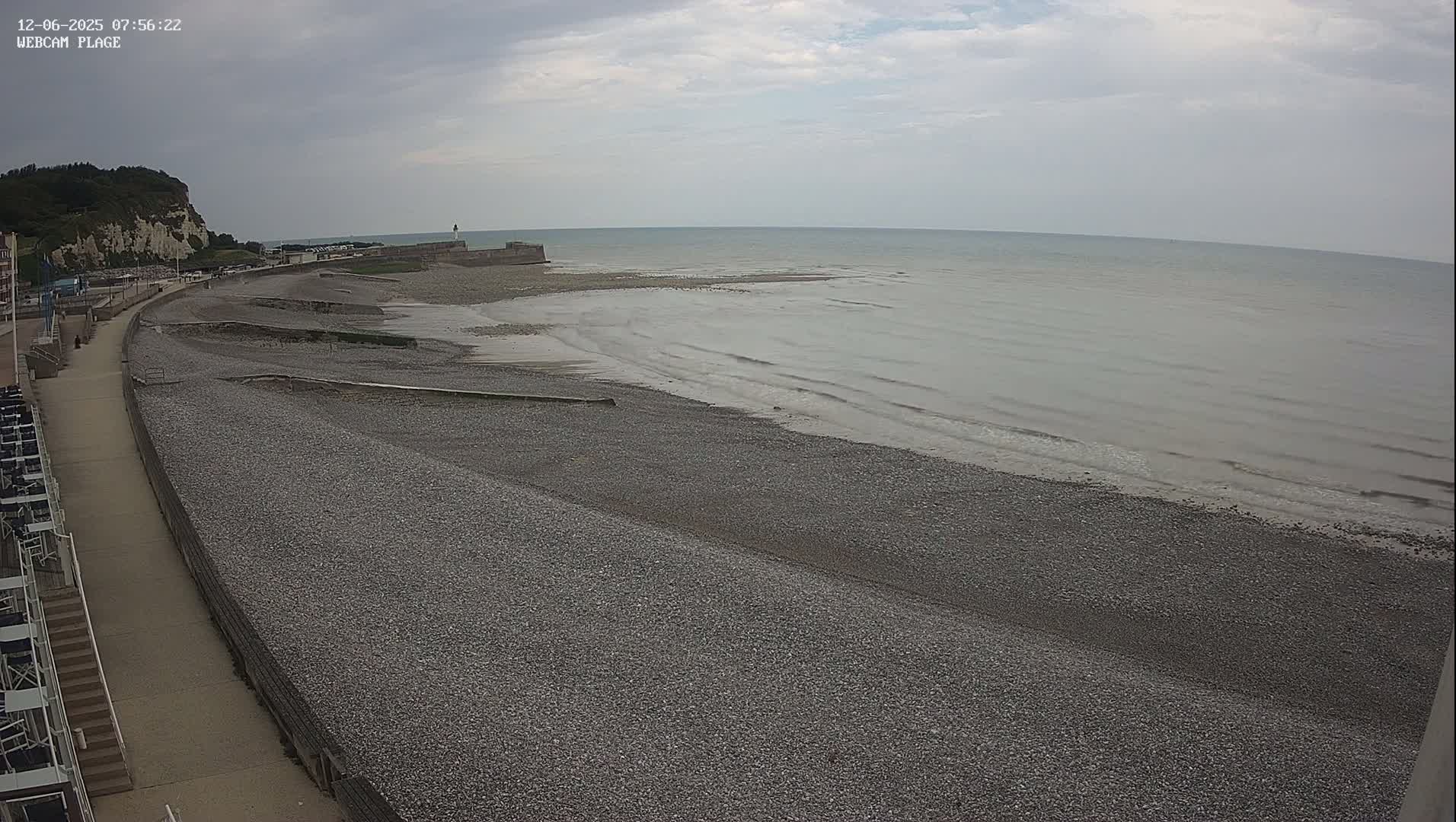 A pebble beach stretches along a calm, gray sea under an overcast sky, with a paved walkway and a cliff visible in the background.