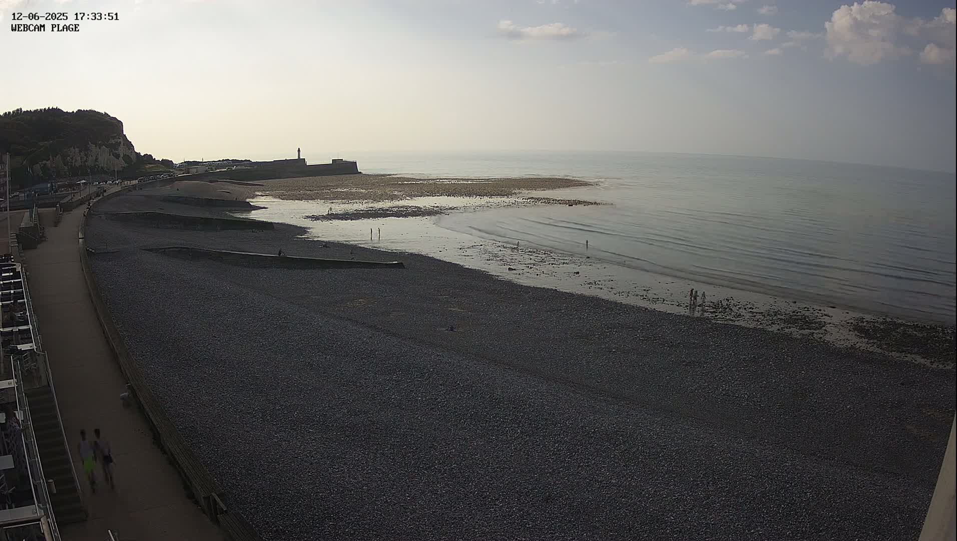 A pebble beach stretches along a calm sea under a partly cloudy sky, with a few people walking along the shore and a paved walkway alongside.
