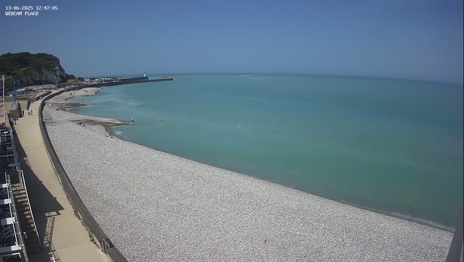 A pebble beach curves gently into a calm, teal sea under a clear blue sky, with a few people scattered along the shore and in the water.