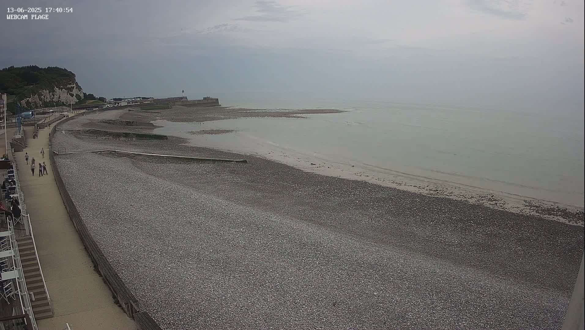 A pebbled beach stretches alongside a paved walkway with several people walking, leading to a calm, light-grey sea under an overcast sky.
