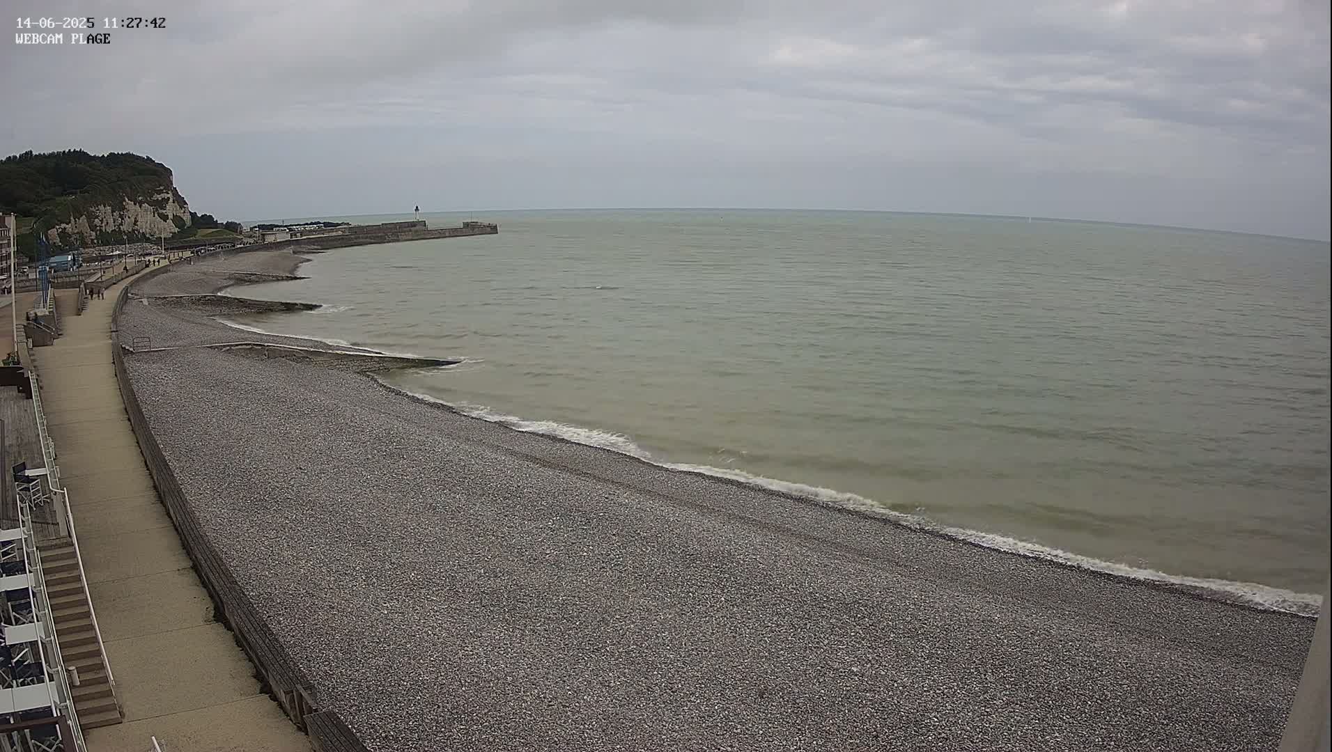 A pebble beach stretches along a calm, gray sea under a cloudy sky, with a paved walkway and a cliff visible in the background.