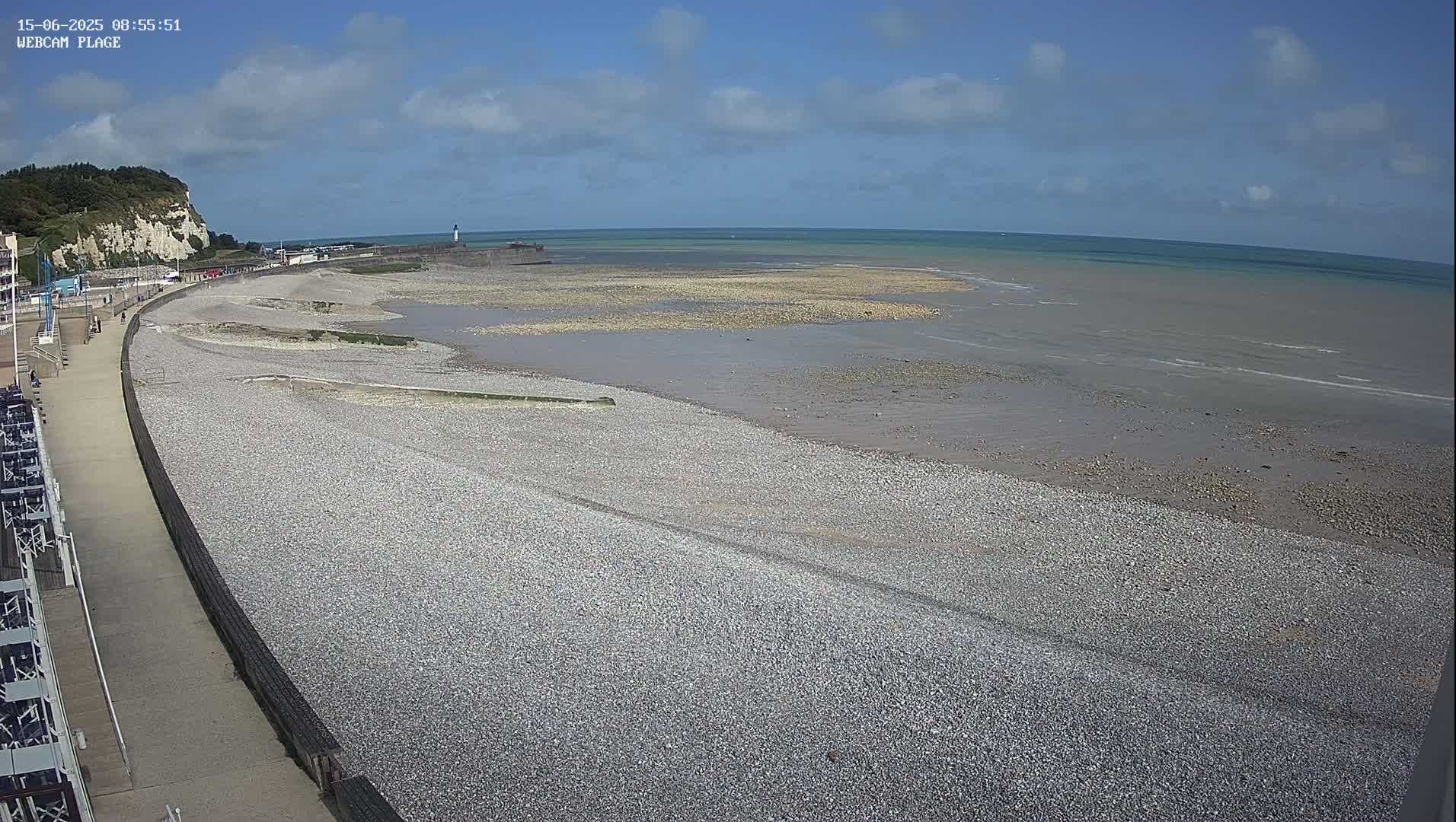 A pebble beach stretches along a paved walkway, meeting a calm sea under a mostly sunny sky, with a white cliff visible in the distance.