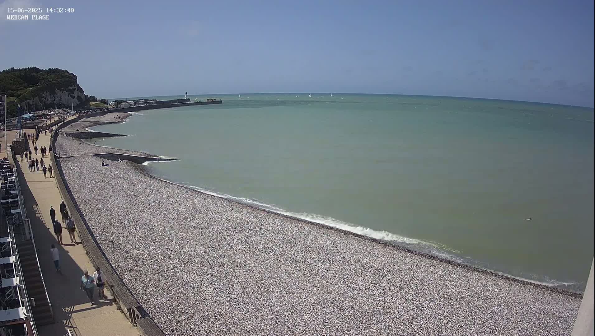 A pebble beach with people walking along a paved path beside it meets a calm, light-green sea under a clear blue sky.