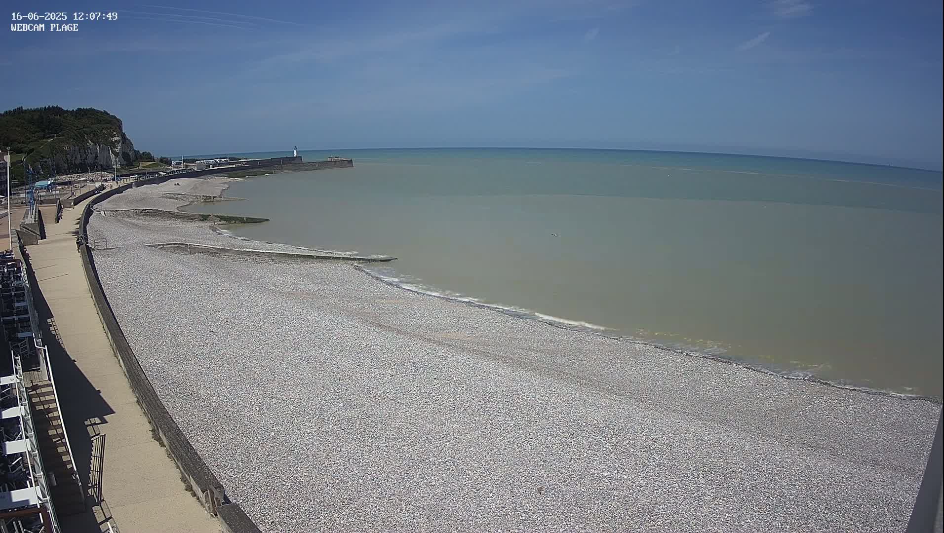 A pebble beach stretches along a calm, light-grey sea under a clear blue sky, next to a paved walkway and a cliff in the background.