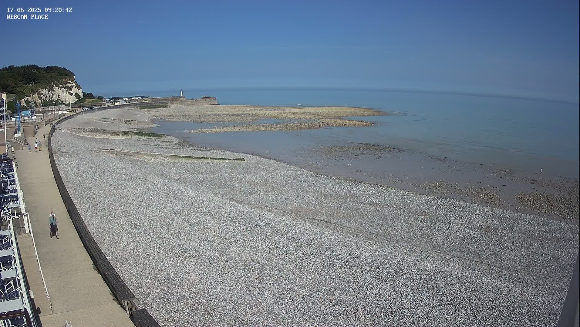A pebble beach stretches toward a calm sea under a clear blue sky, with a promenade and a few people visible along the shoreline.