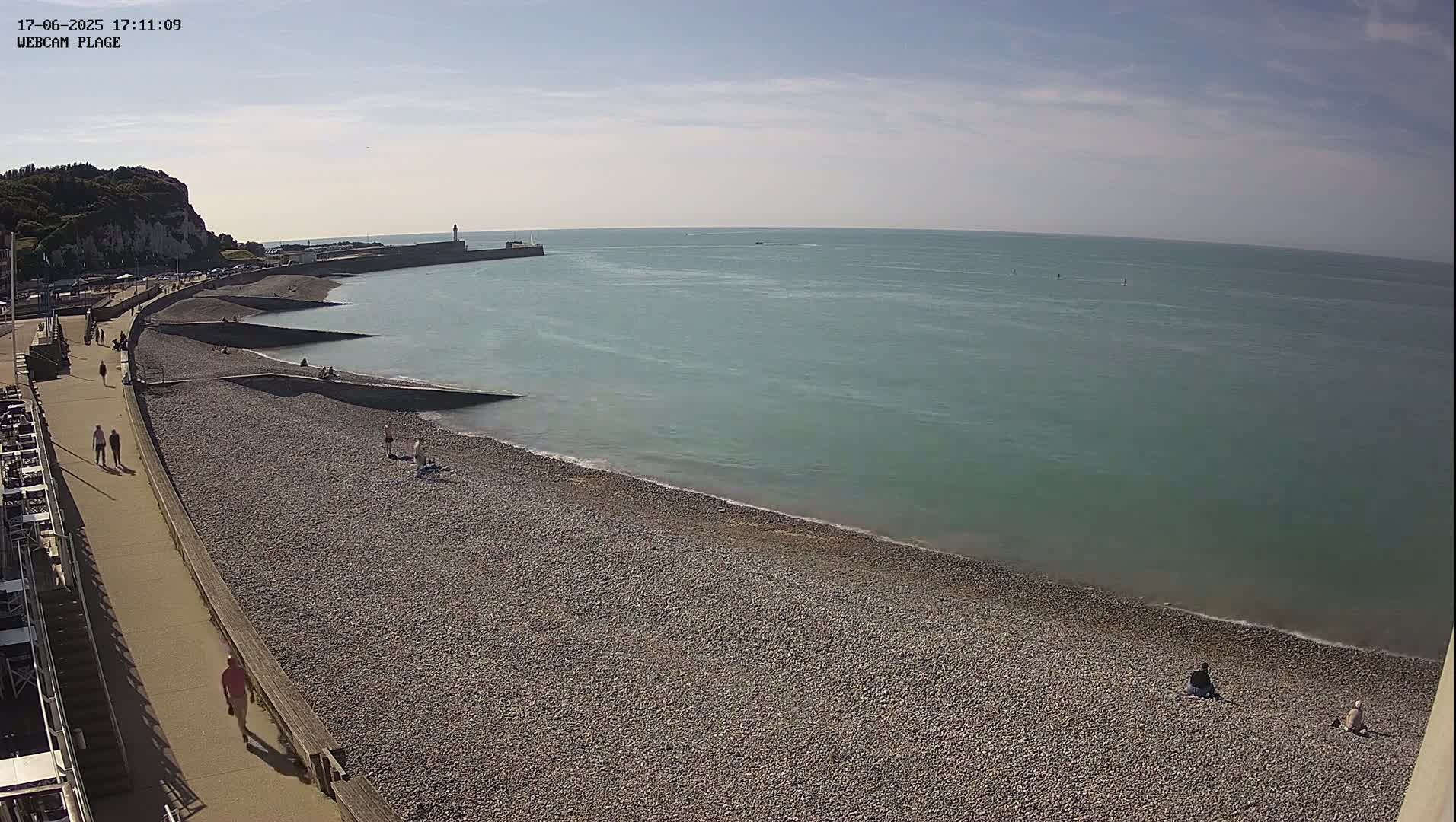 A pebble beach stretches along a calm, teal ocean under a mostly sunny sky, with a few people scattered along the shore and a walkway with some people walking along it.