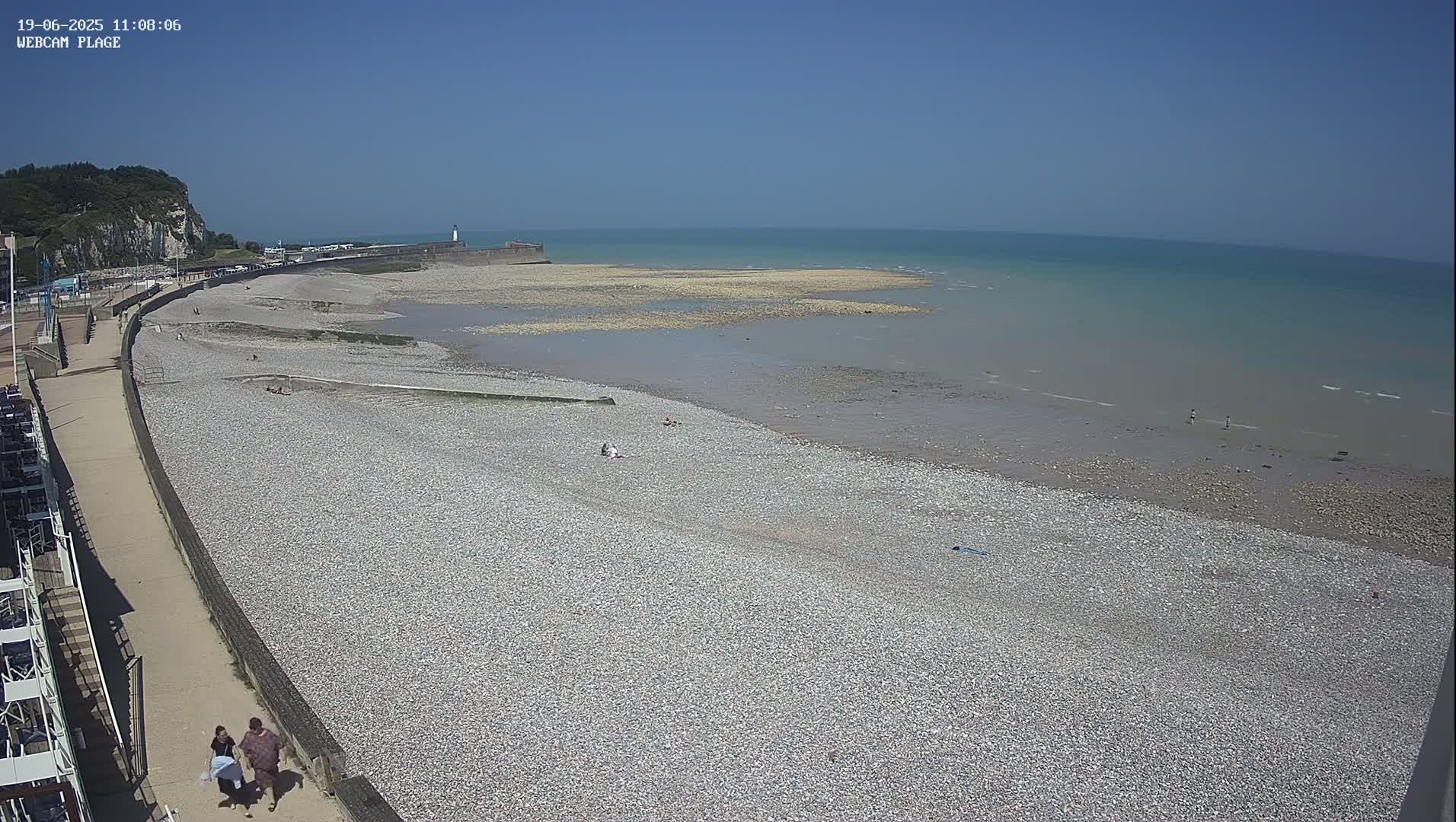 A pebble beach stretches out to a calm, light-blue sea under a clear blue sky, with a few people visible on the beach and a walkway along the shore.