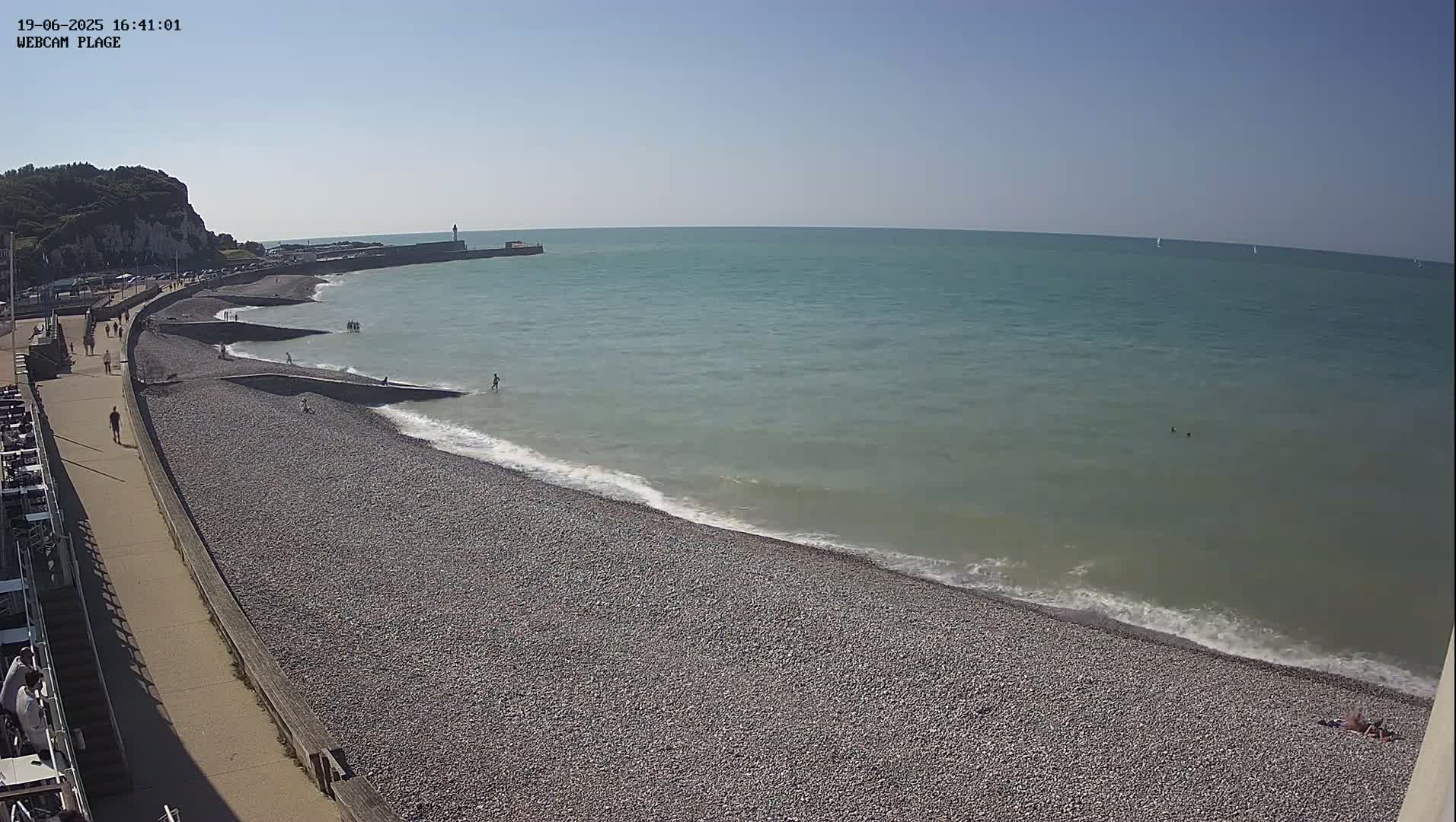 A pebble beach with a few people on it and in the water stretches along a calm, teal ocean under a clear, sunny sky.