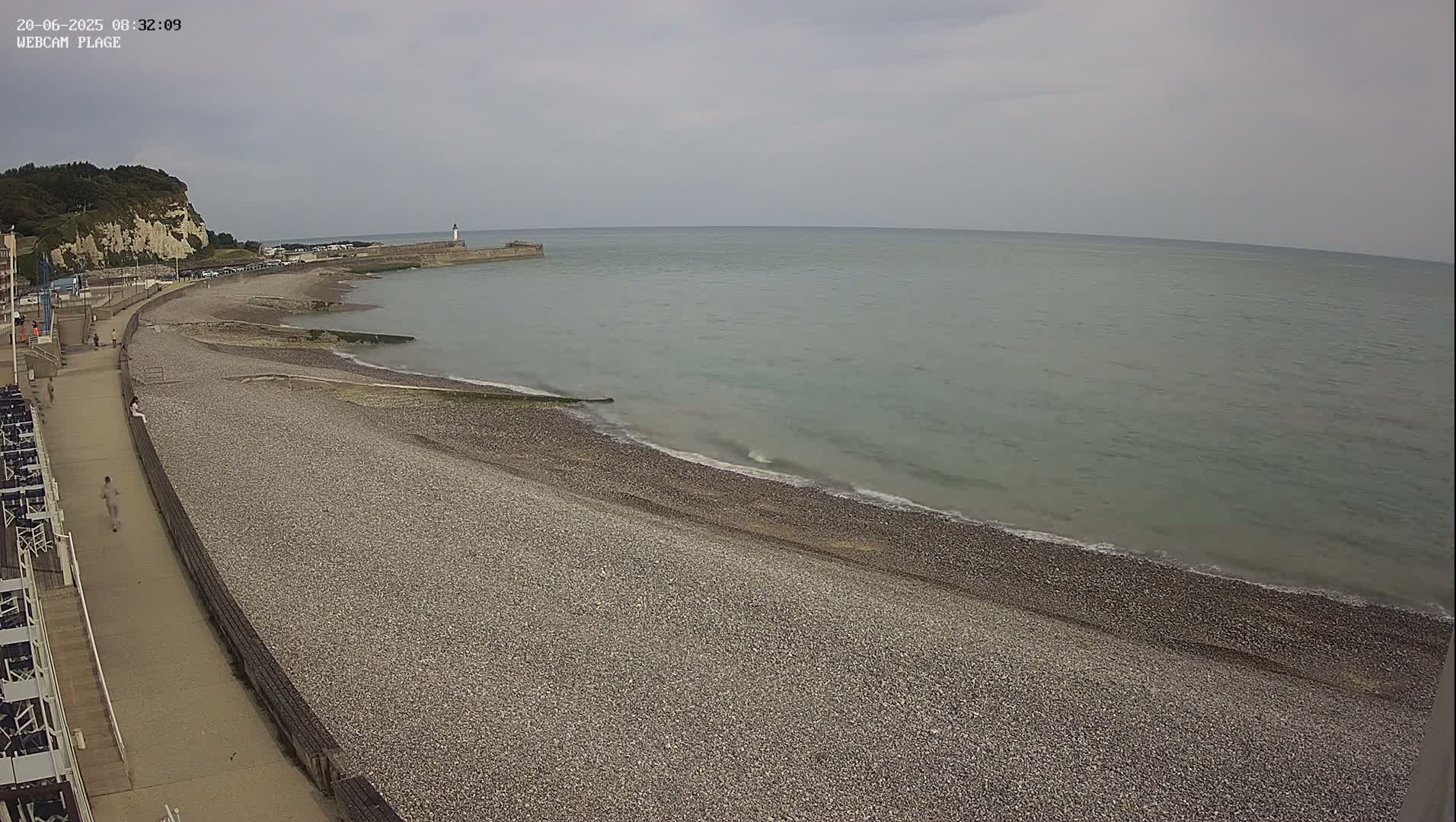 A pebbled beach stretches along a calm, gray-green sea under an overcast sky, with a walkway and a lighthouse visible in the distance.