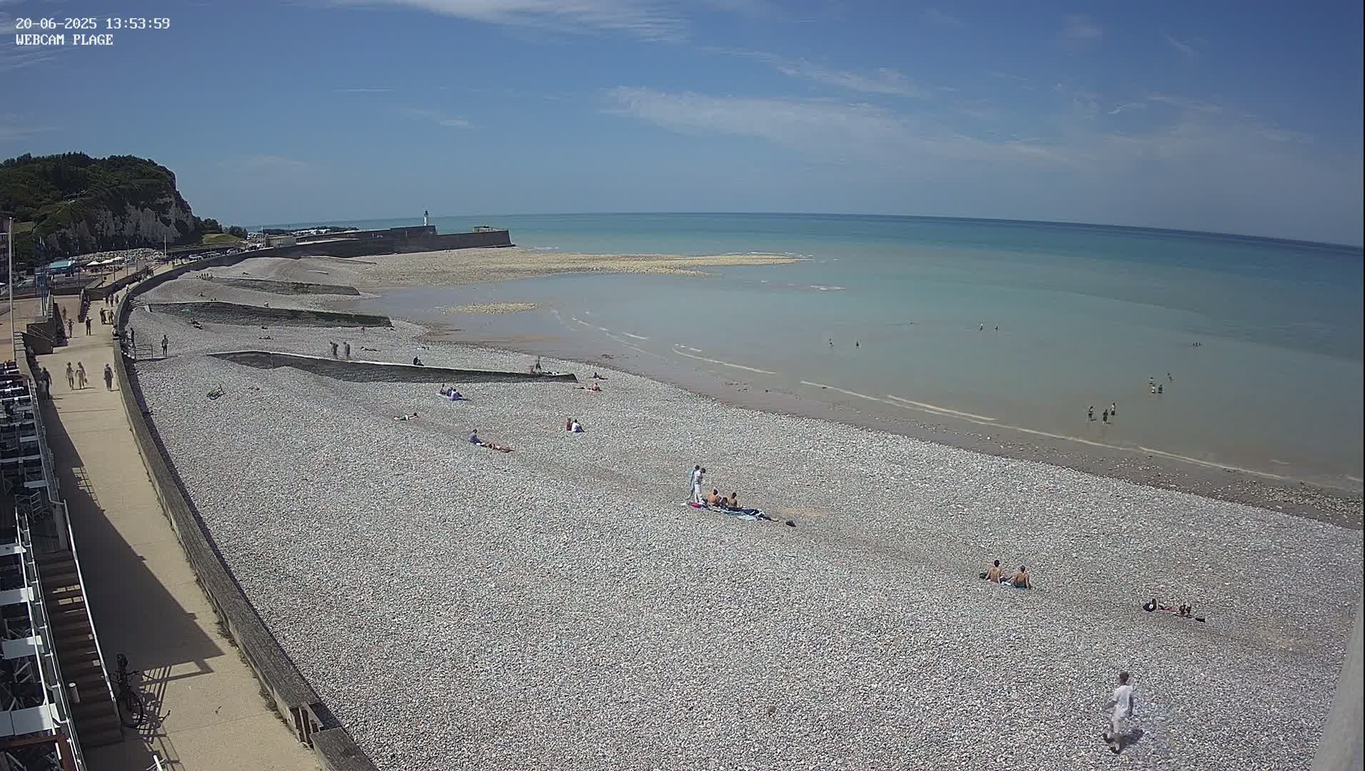 A pebble beach with scattered sunbathers is shown, next to a calm, light-blue ocean under a clear, sunny sky.
