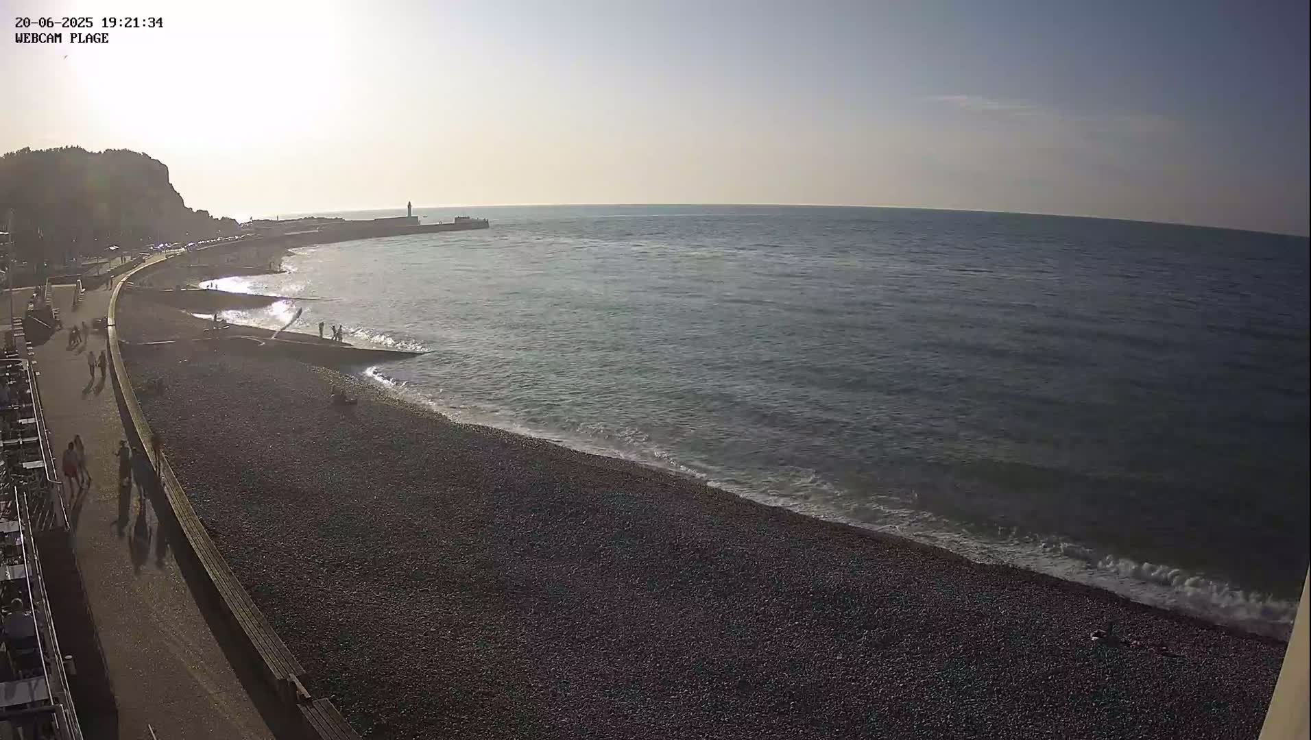 A pebble beach with a few people walking along a paved path next to it, under a mostly clear sky, meets a calm sea with small waves lapping the shore.