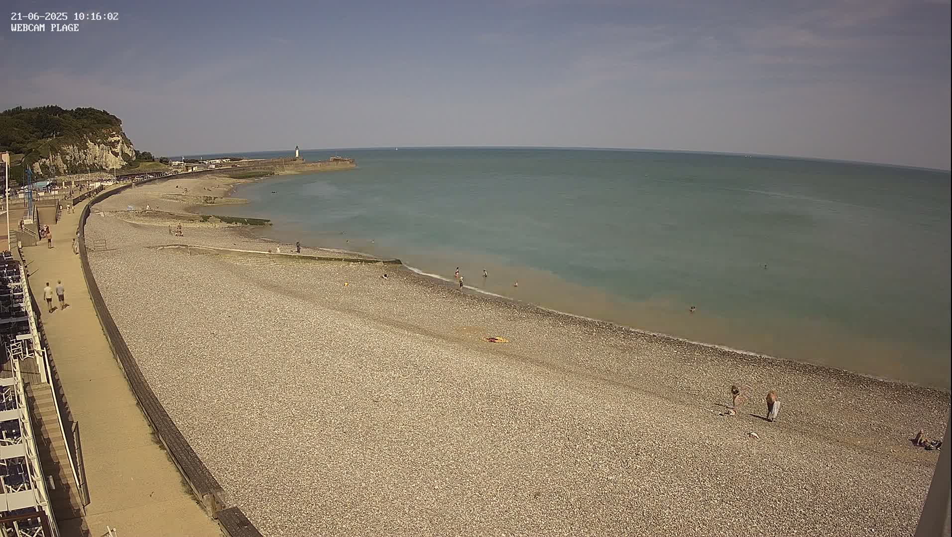 A pebble beach stretches along a calm, light-green sea under a clear, sunny sky, with a few people swimming and walking on the shore and along a paved walkway.