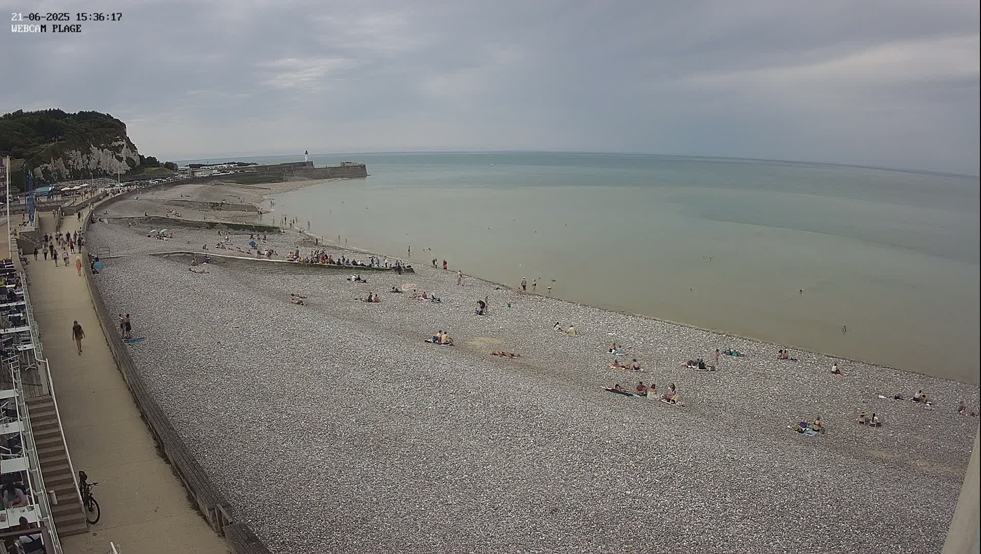 A pebble beach with numerous sunbathers and a few people swimming in a calm, light-grey sea under an overcast sky.