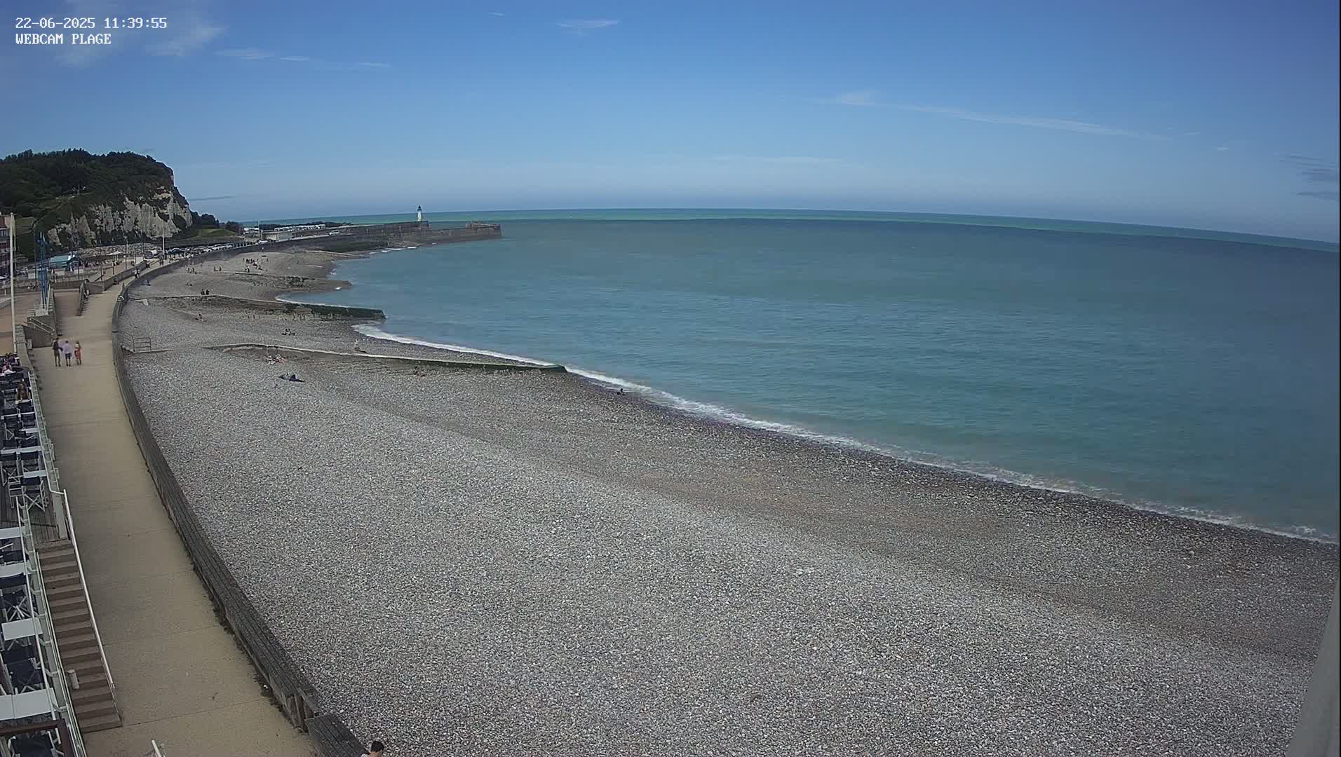 A pebble beach stretches along a calm ocean under a clear blue sky, with a walkway and a few people visible.