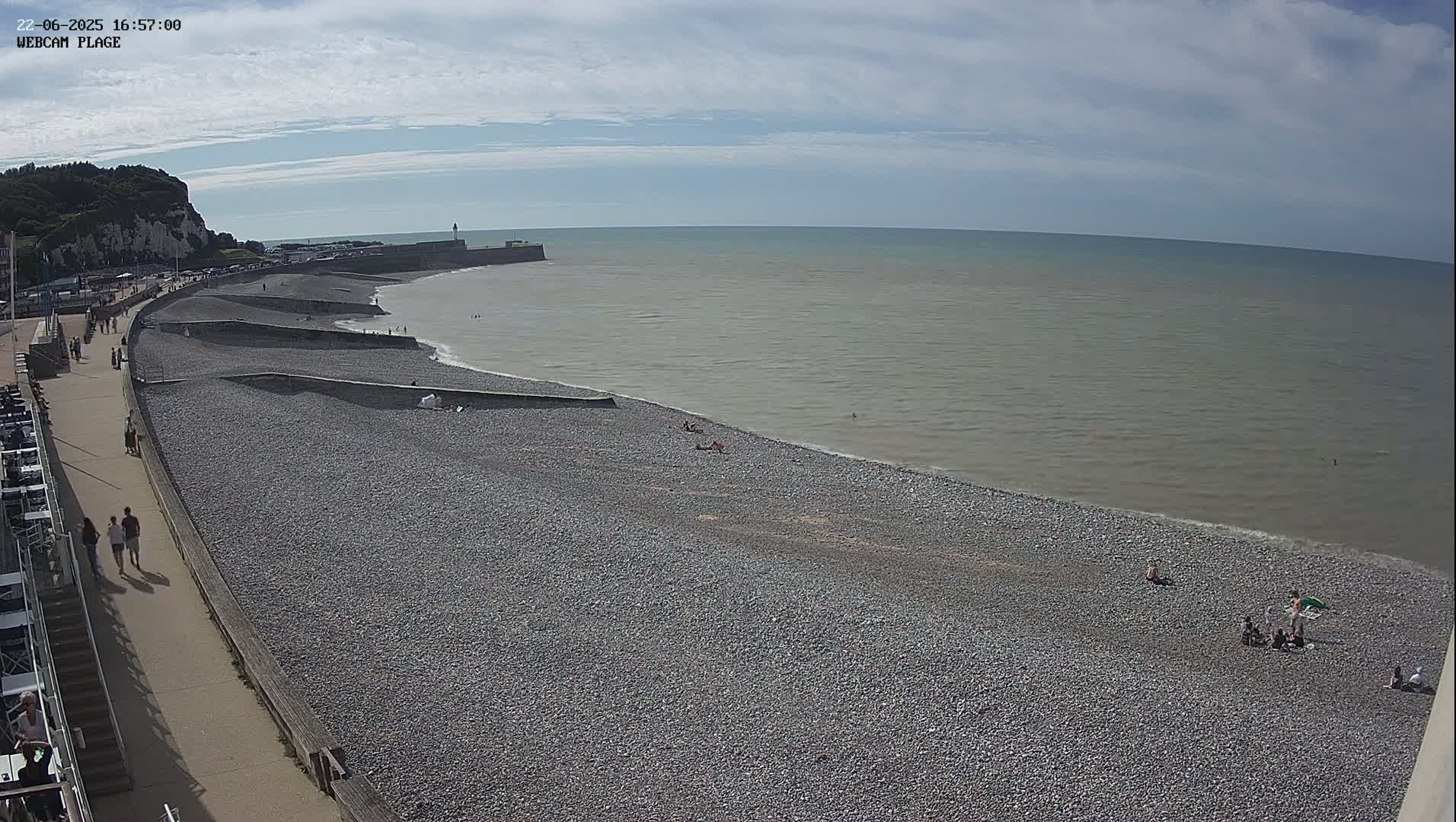 A pebble beach with a few people sunbathing and walking along a promenade, under a partly cloudy sky.