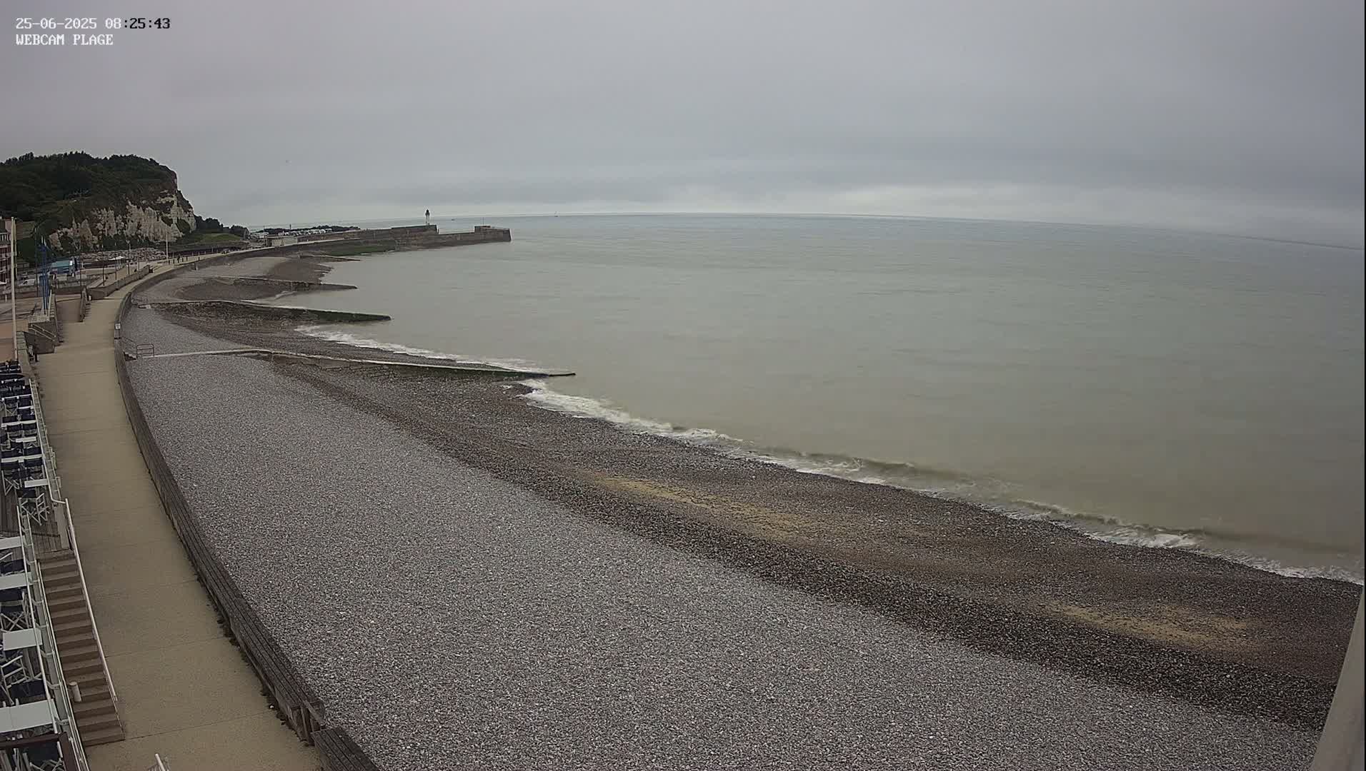 A pebble beach stretches along a calm, gray sea under an overcast sky, next to a paved walkway and a cliff in the background.