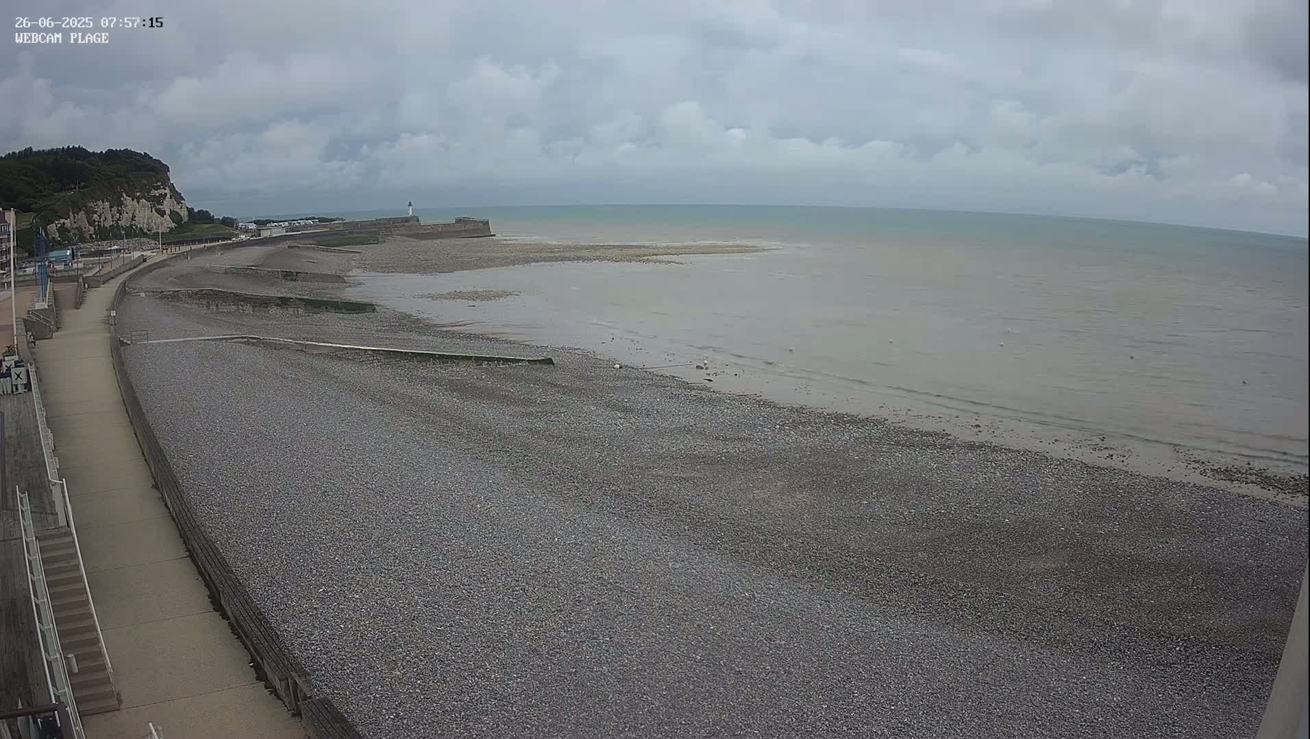 A pebble beach stretches along a calm, gray sea under a cloudy sky, with a paved walkway and a cliff visible in the background.