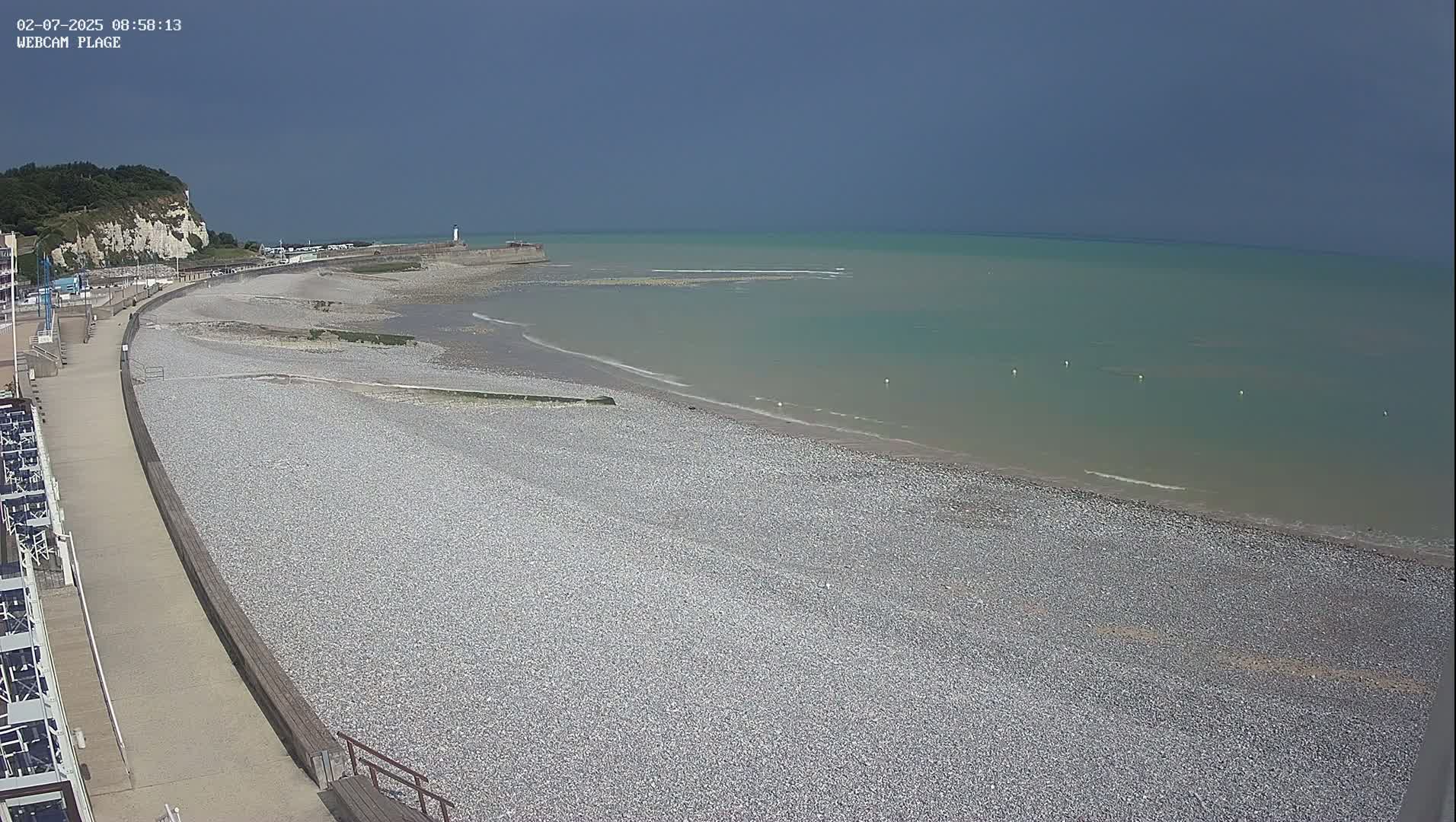 A pebble beach curves along a calm, light-green sea under a mostly clear, grayish-blue sky, with a paved walkway and buildings visible along the shore.