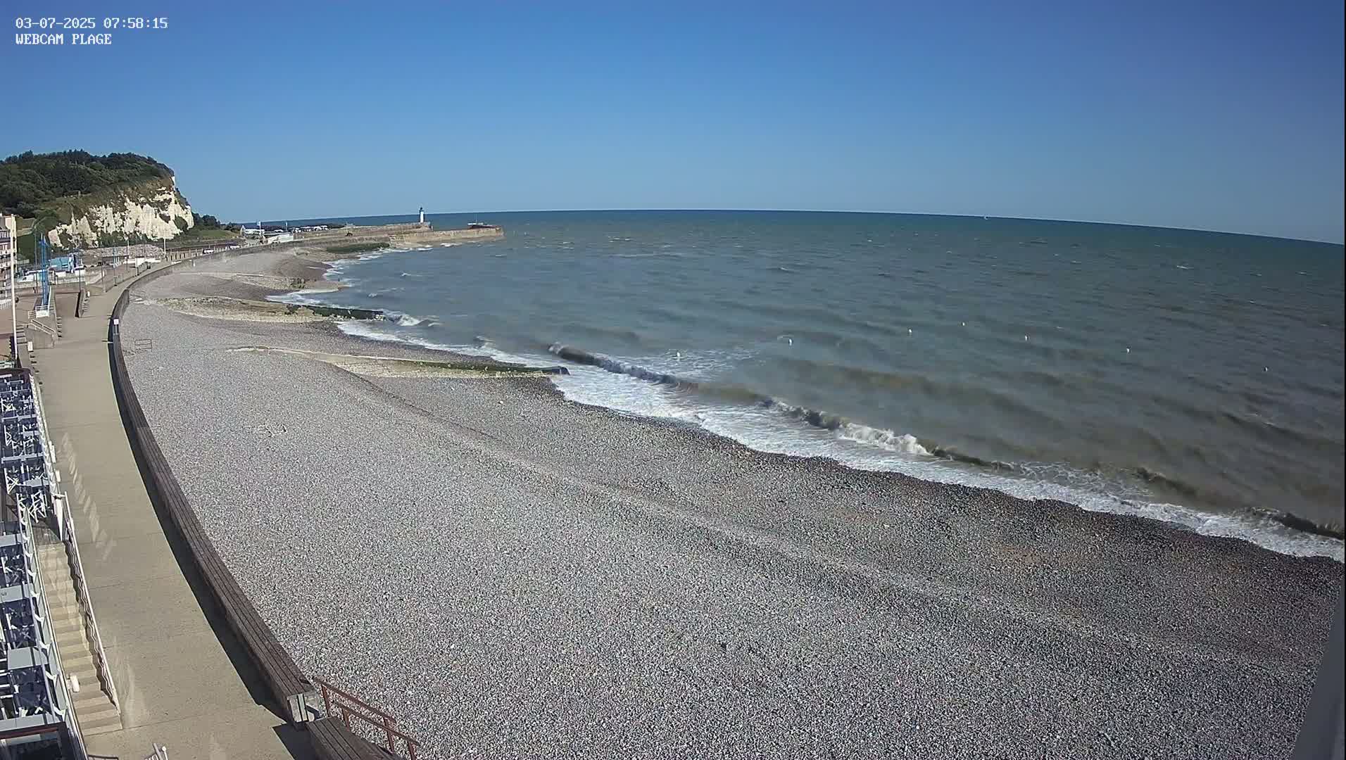 A pebble beach stretches along a calm sea under a clear blue sky, with a walkway and white cliffs visible in the background.