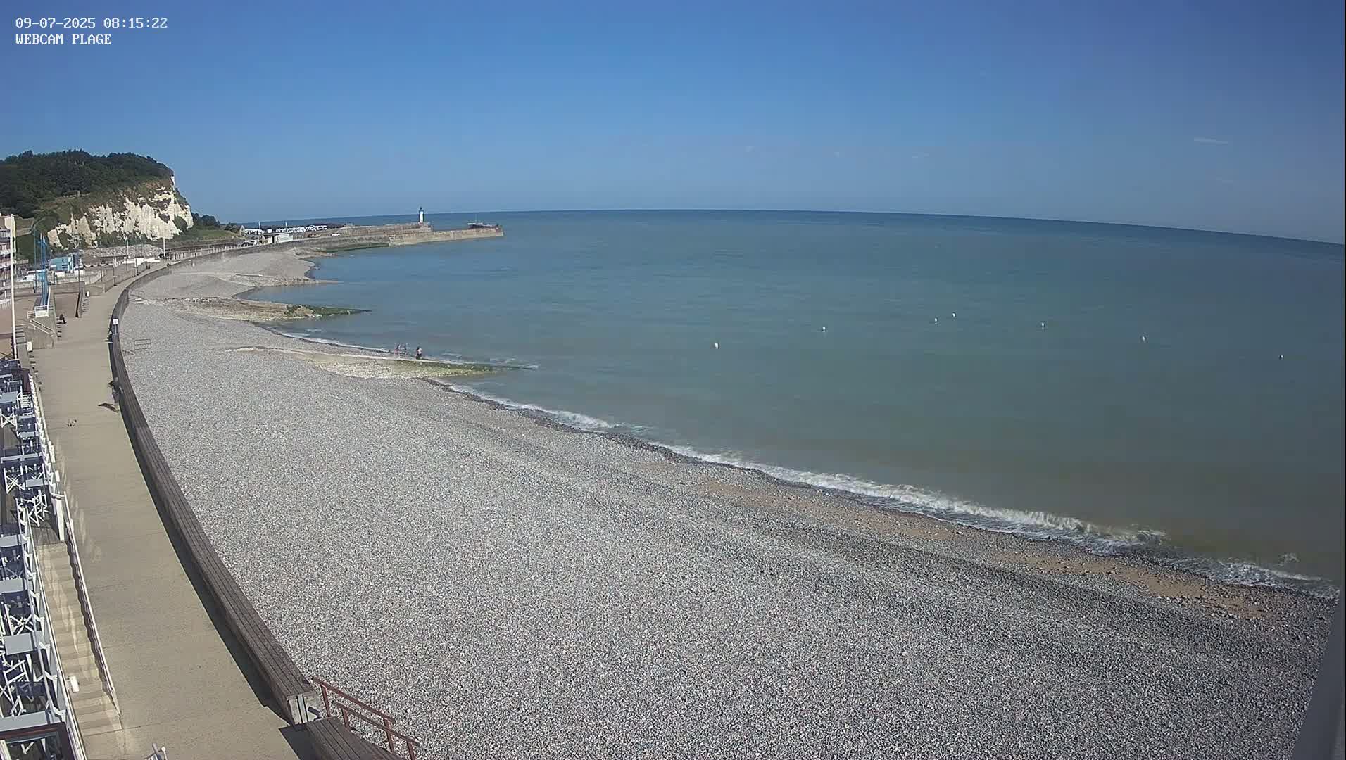 A pebble beach stretches along a calm sea under a clear blue sky, with a walkway and white cliffs visible in the background.
