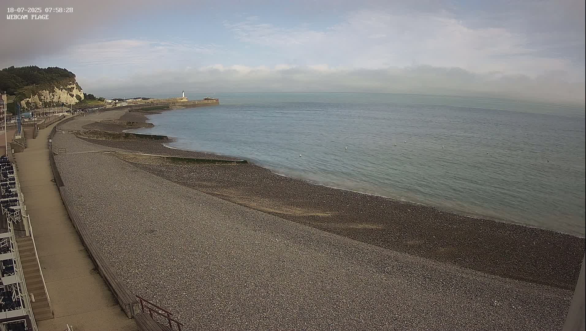 A pebble beach stretches alongside a paved walkway, leading to a calm sea under a mostly cloudy sky, with a distant lighthouse and cliff visible.