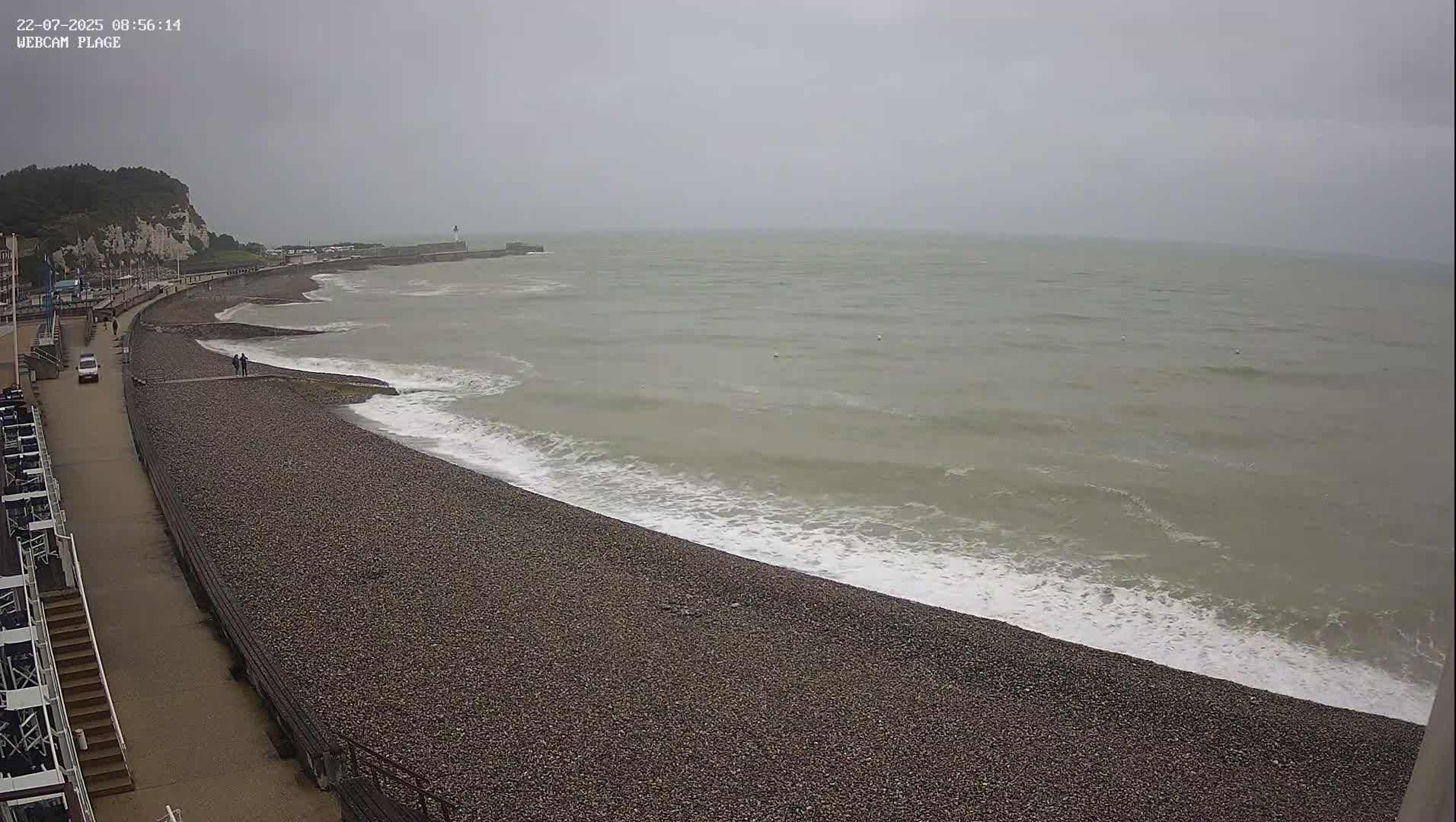 A pebble beach stretches along a gray, overcast sea, with a walkway and white buildings visible in the background.